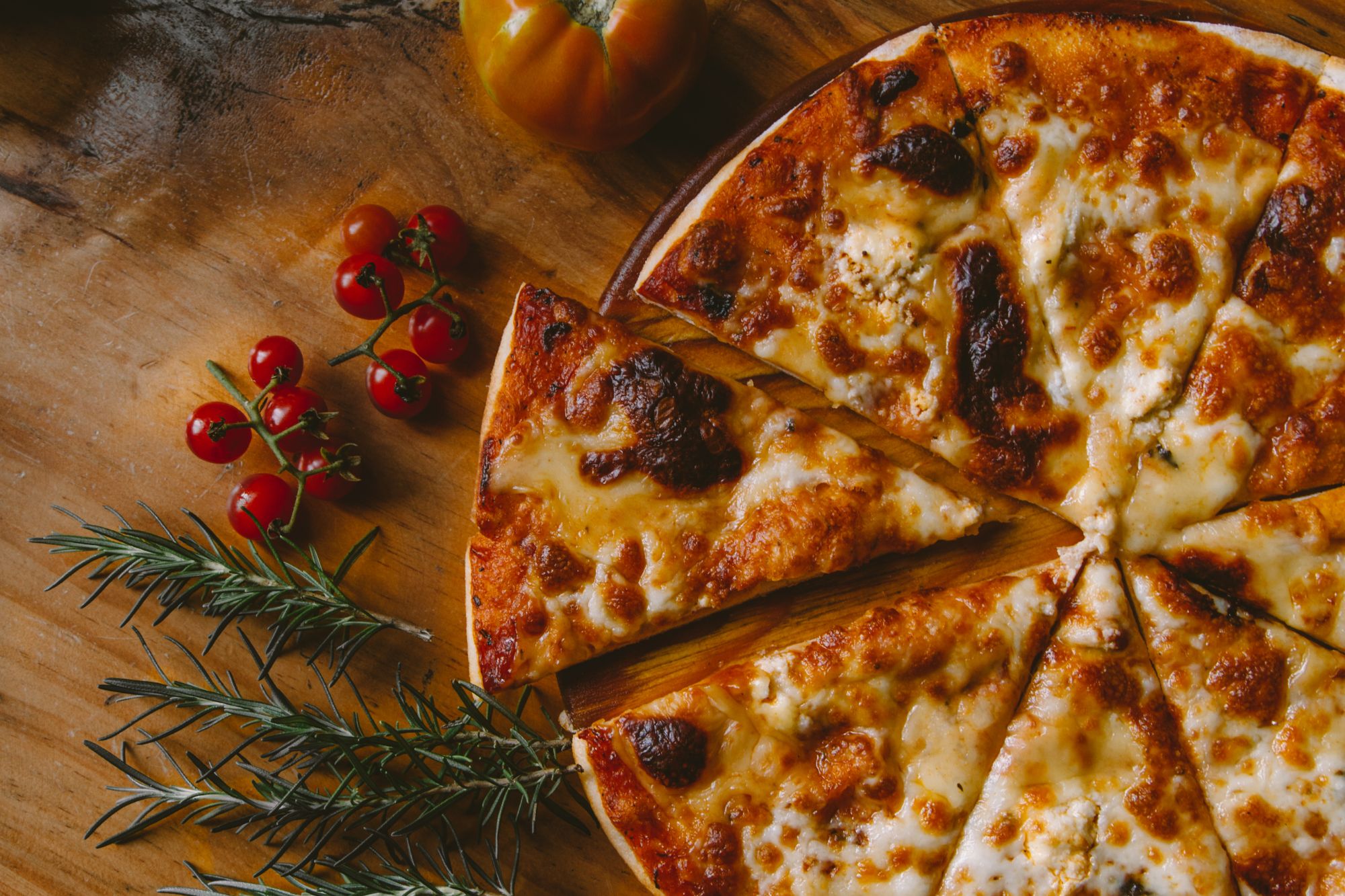 Półmisek pizzy z serami na drewnianym stole, obok pomidory i gałązki rozmarynu.