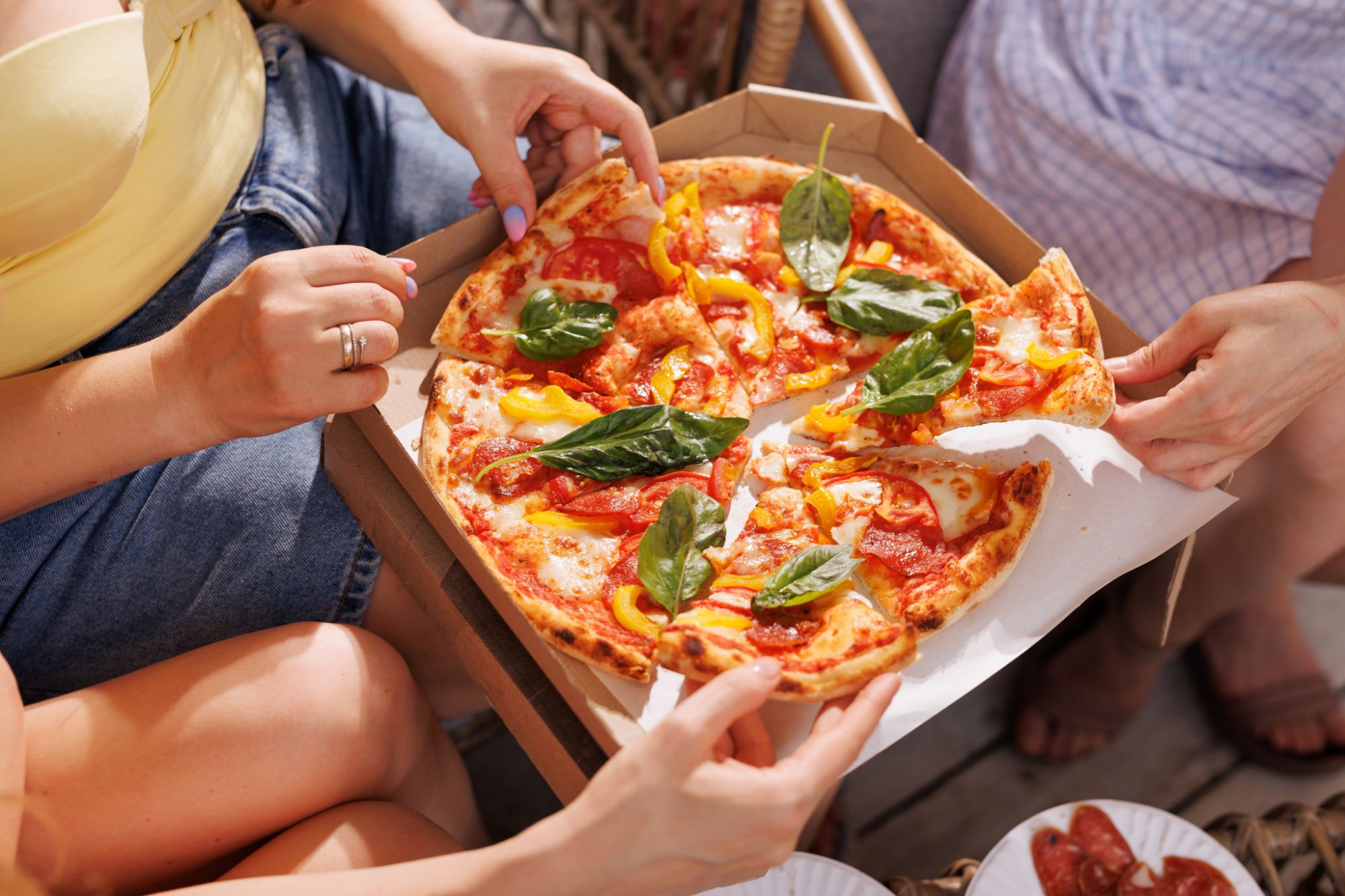 Dwie osoby siedzące na zewnątrz trzymają pizzę z serem, sosem pomidorowym i świeżymi warzywami.