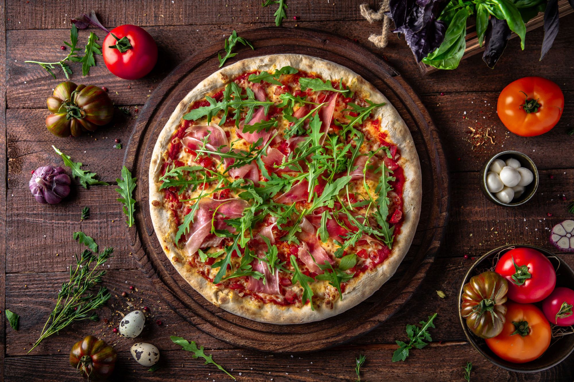 Pizze s paradajkami, šunkou a rukolou na drevenej doske so zeleninou okolo.