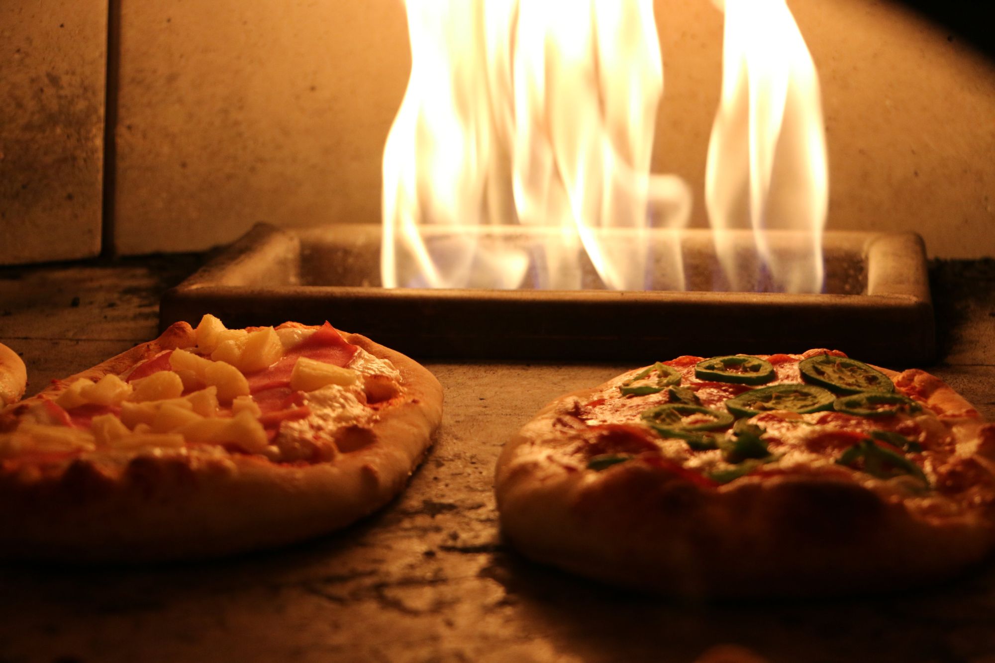 Dve pizze sa pečú v peci, jedna s ananásom a šunkou, druhá so zelenou paprikou a omáčkou.