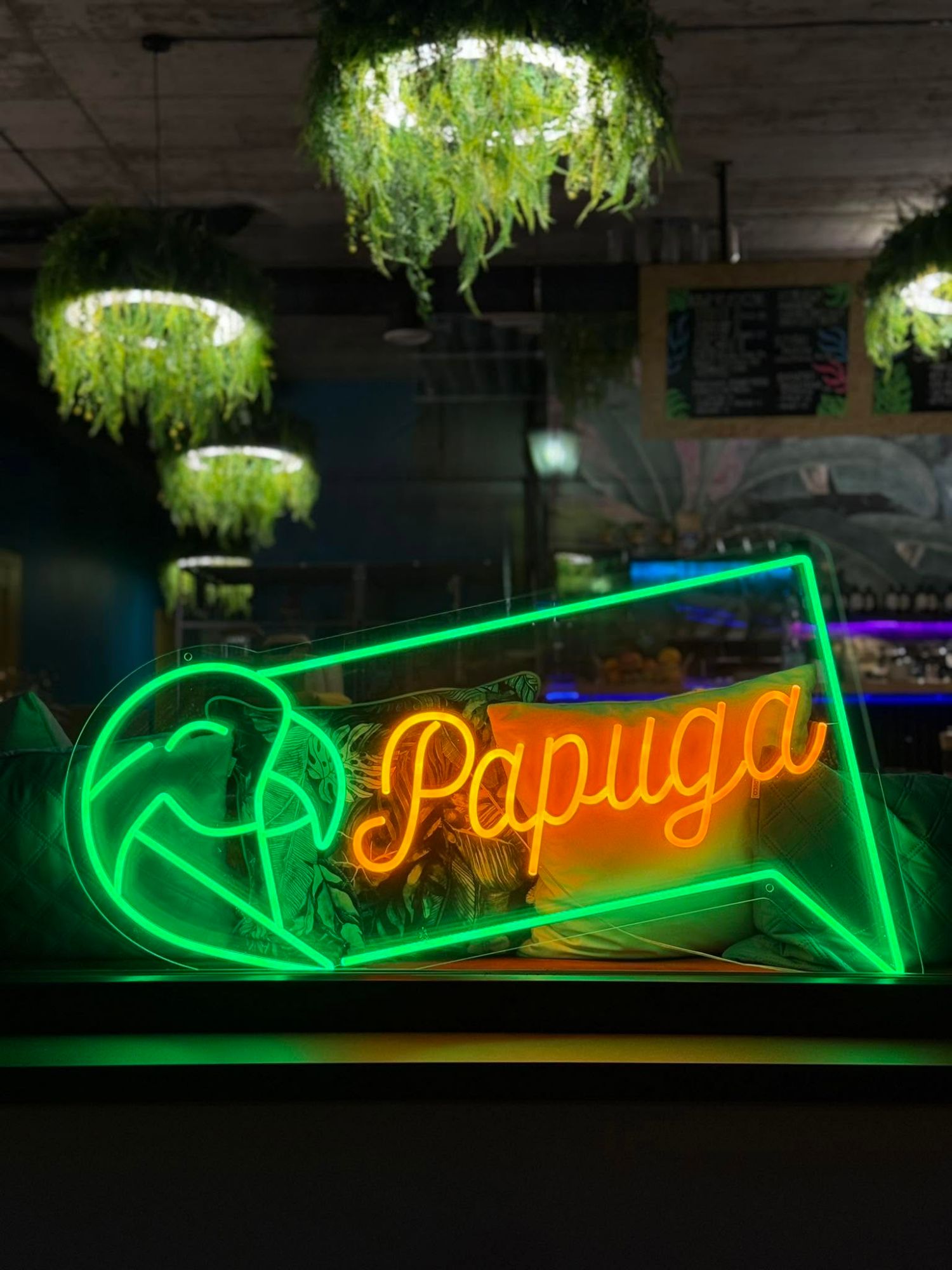 Neonowy znak z napisem "Papuga" na tle zielonych roślin w nowoczesnej restauracji.