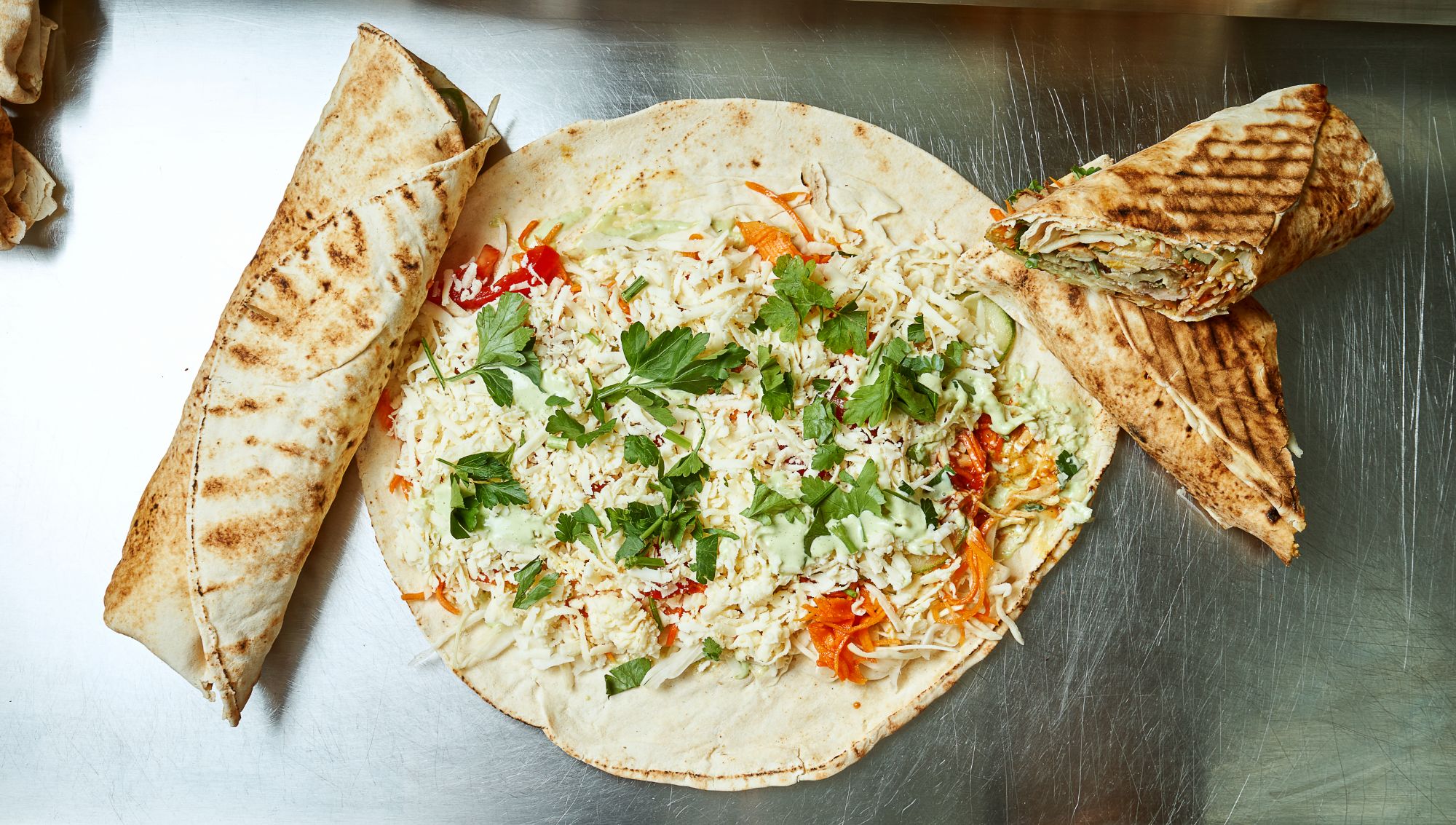 Tortilla wypełniona sałatką, ziołami i serem, obok dwa zrolowane burrito.