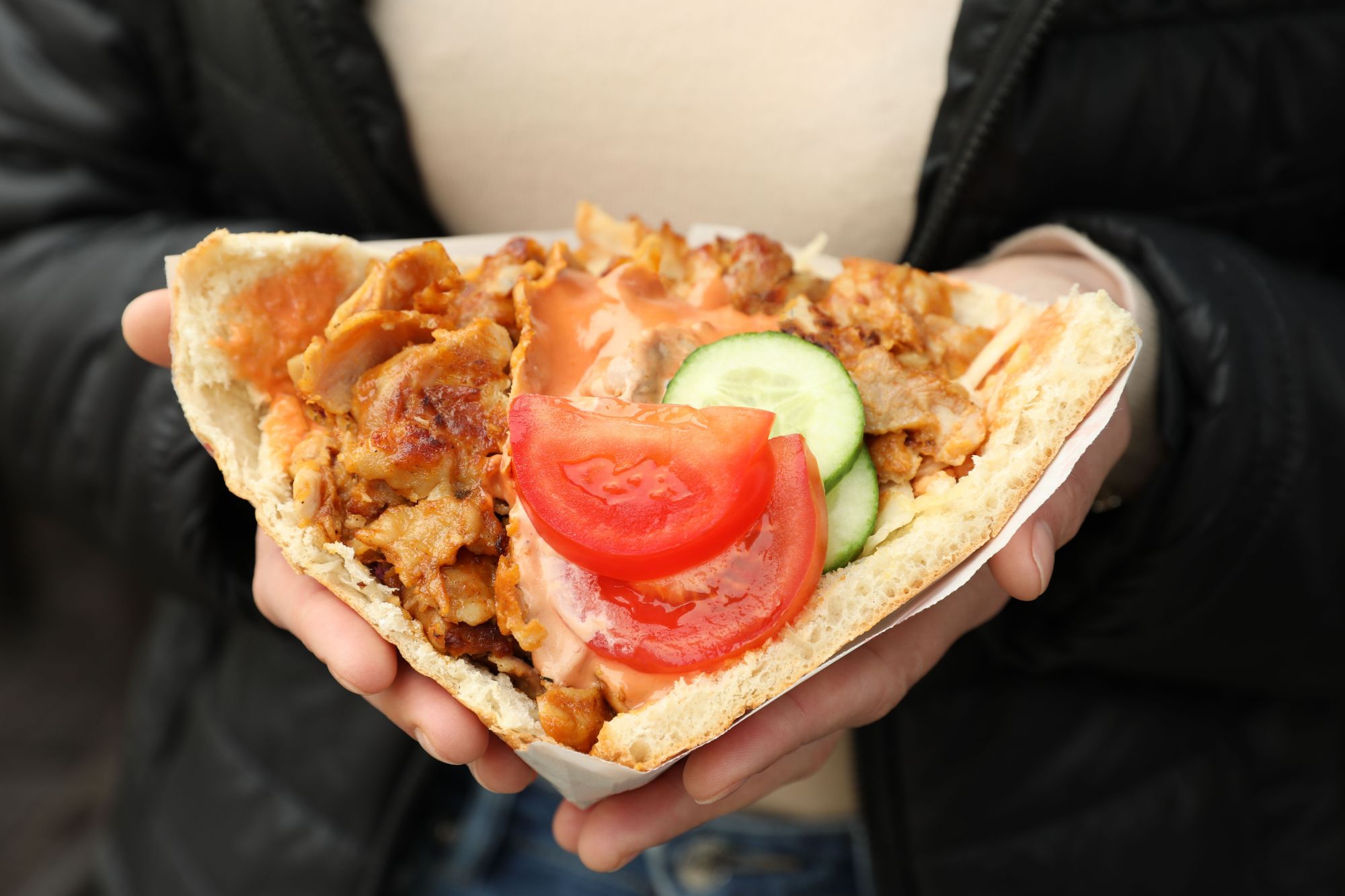 Kanapka z kurczakiem, pomidorem i ogórkiem w pita, trzymana w dłoniach na zewnątrz.