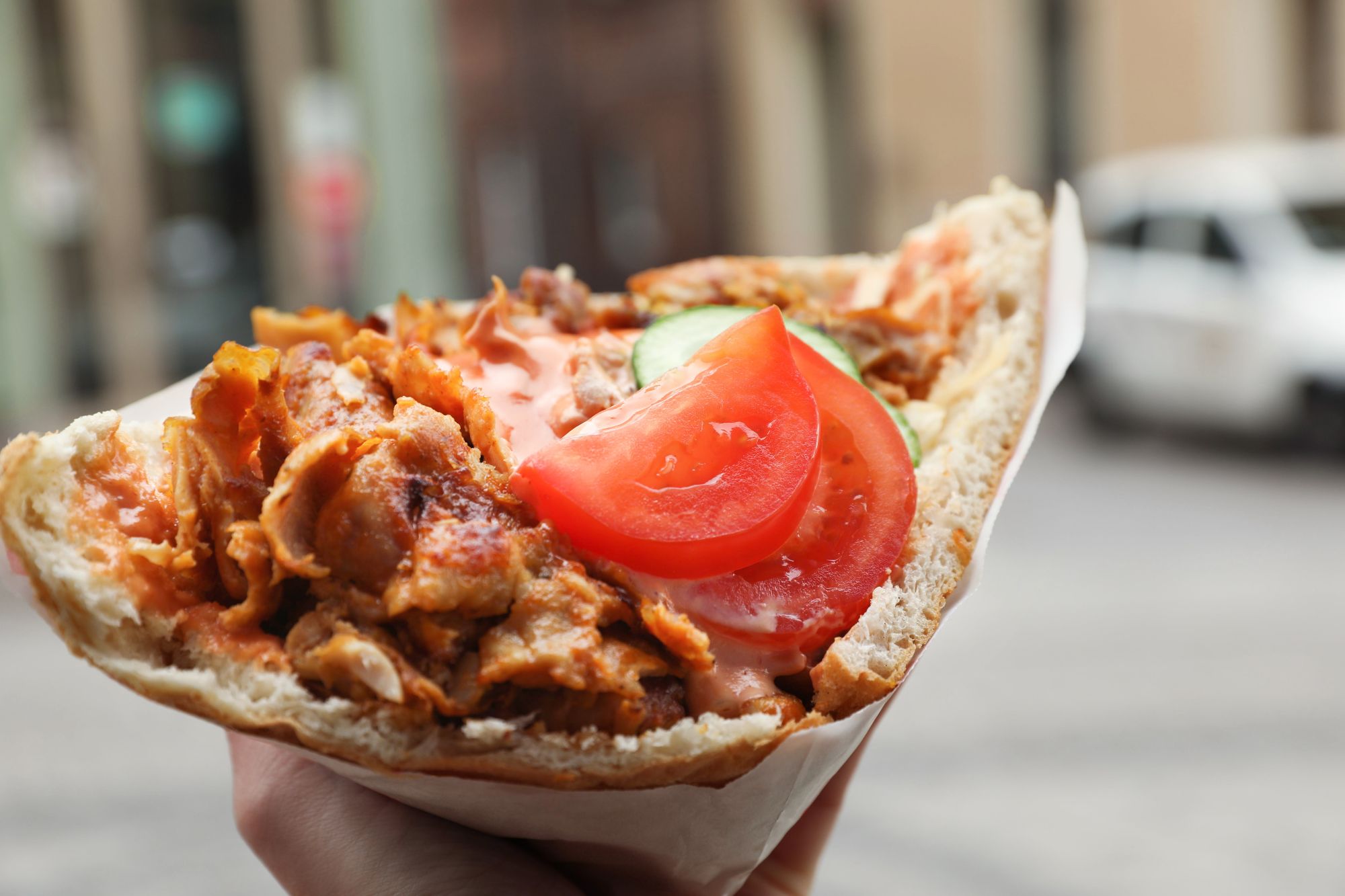 Kebab z kurczaka w chlebie pita z pomidorami i ogórkiem, trzymany w ręku na ulicy.