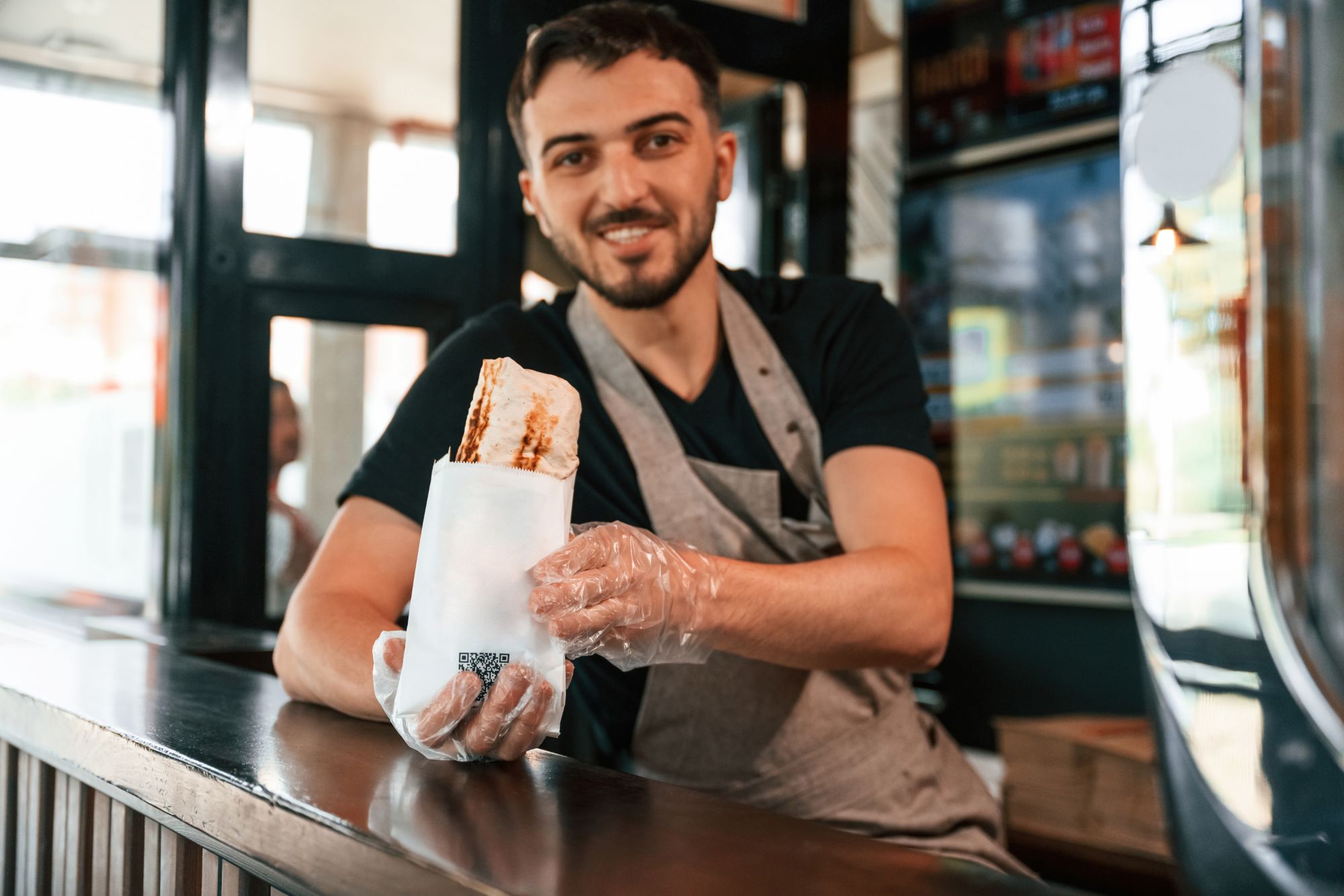 Mężczyzna w fartuchu trzyma kanapkę w papierze, uśmiechając się przy ladzie w food trucku.