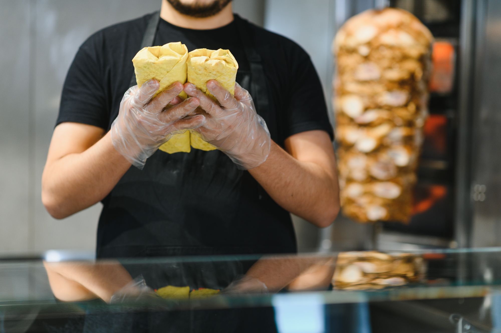 Osoba trzymająca dwa zawiniątka w barze szybkiej obsługi, w tle obracający się kebab.