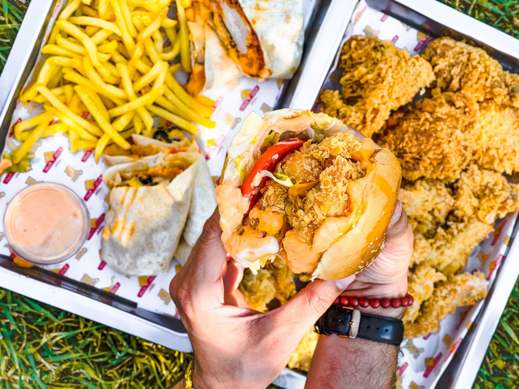 Osoba trzyma burgera z kurczakiem, obok frytki i kawałki smażonego kurczaka na talerzu.