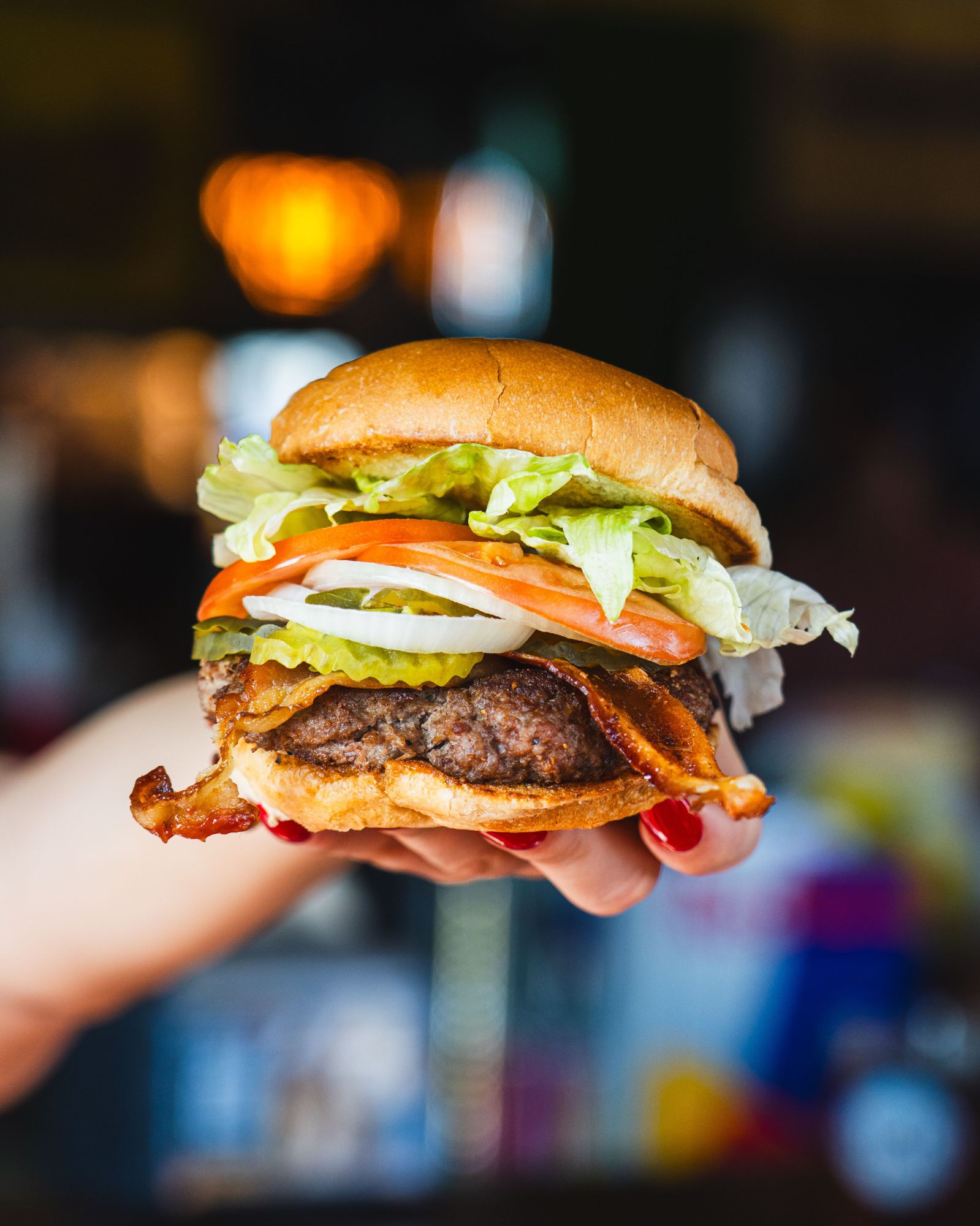 Burger z soczystym mięsem, sałatą, pomidorem, cebulą i ogórkiem, trzymany w ręku na tle rozmytej restauracji.