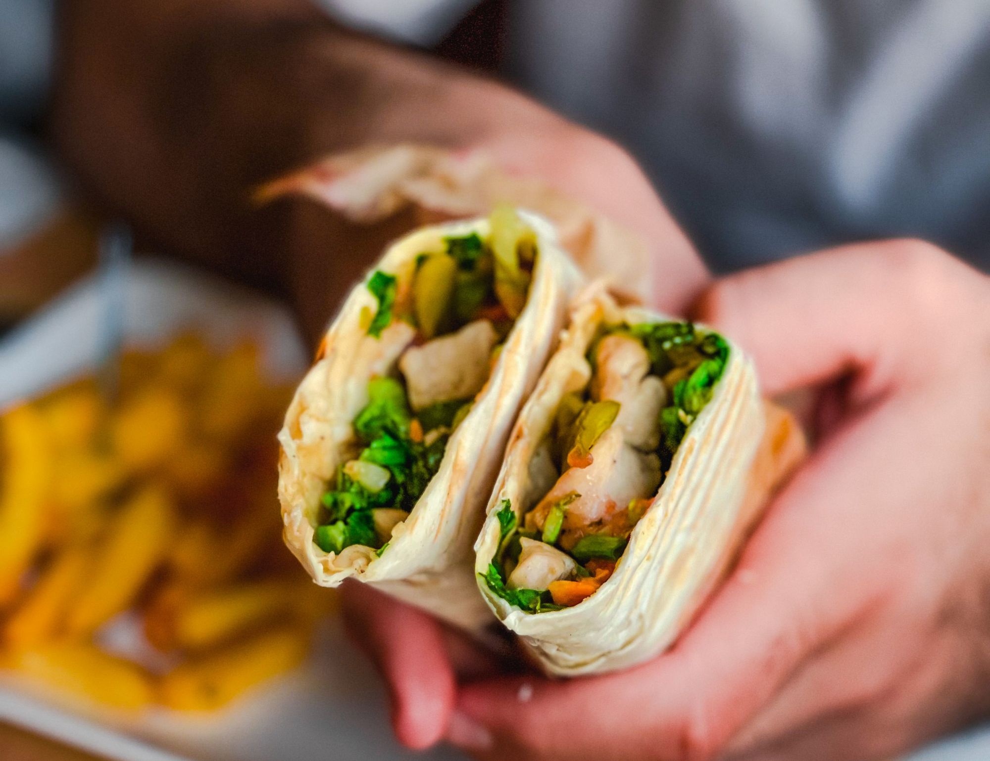 Osoba trzyma burrito z drobiem, sałatą i warzywami, obok frytki na talerzu.