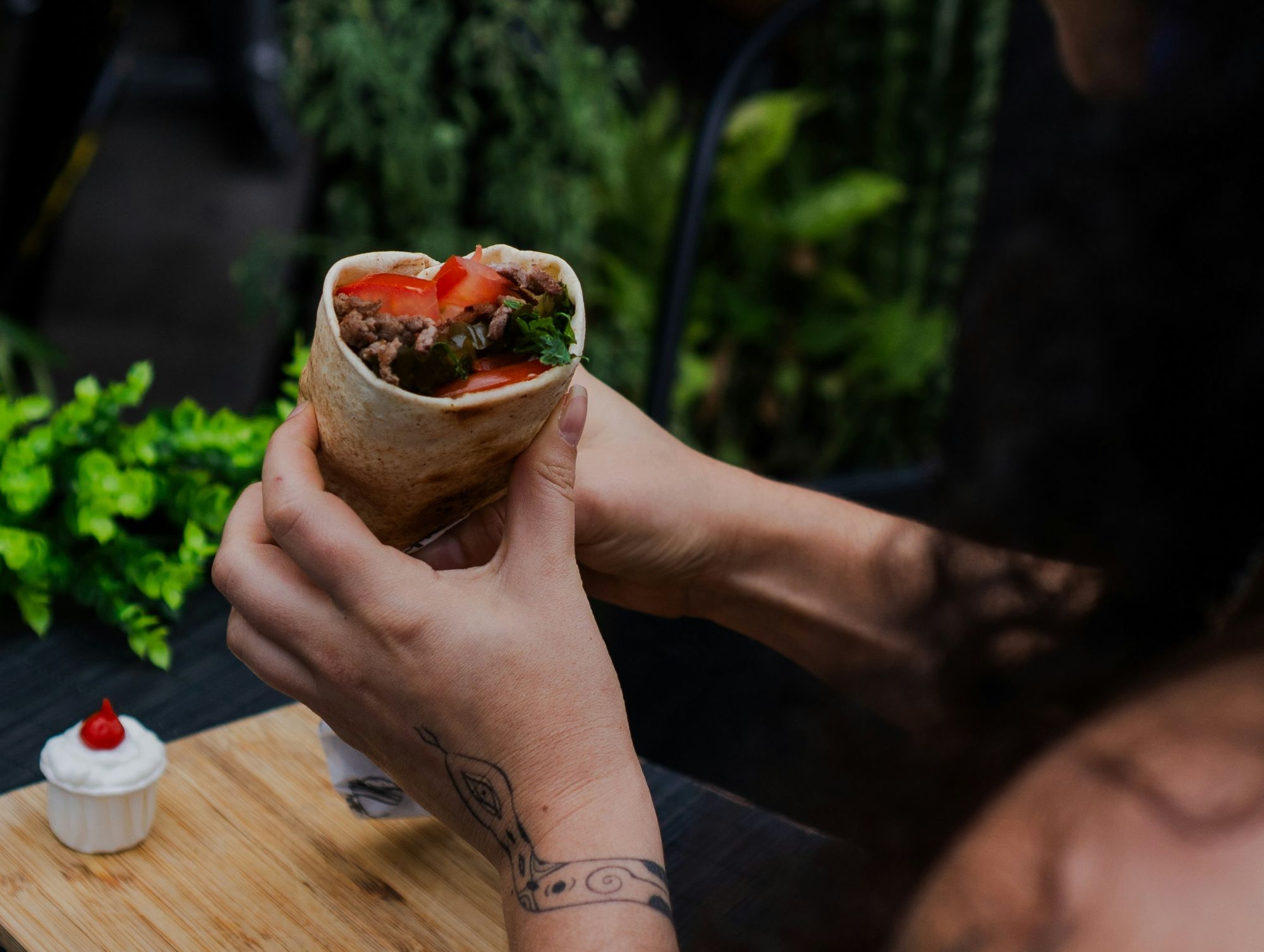 Osoba trzymająca burrito z warzywami w otoczeniu roślin, obok mini deser w filiżance.