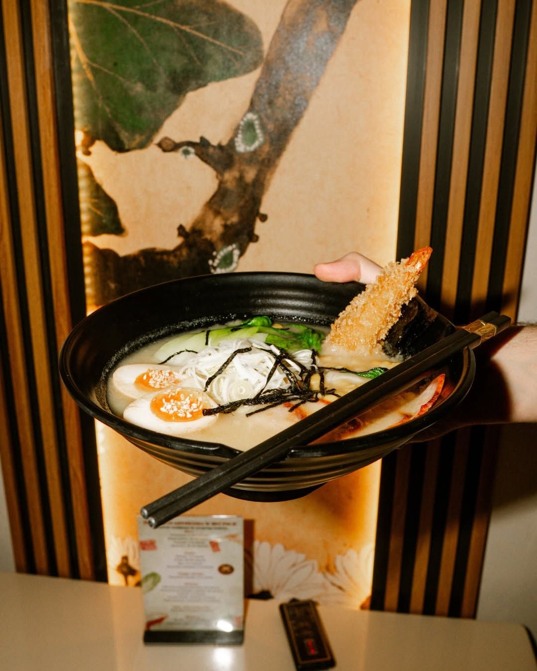 Osoba trzyma misę ramen z makaronem, jajkiem, wodorostami i chrupiącym kawałkiem ryby, na tle dekoracyjnej ściany.