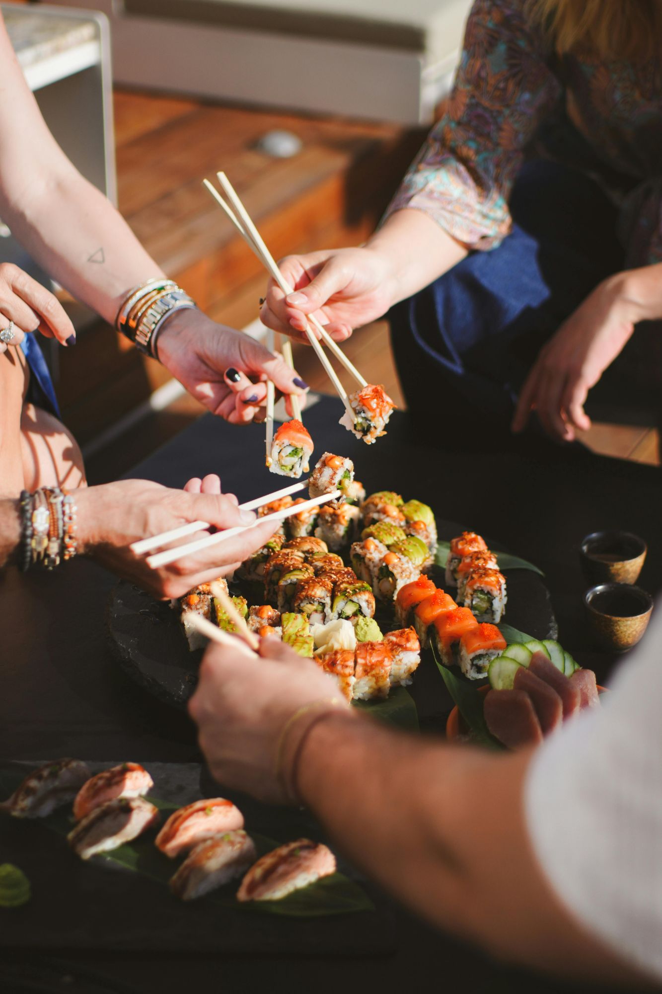 Grupa ludzi dzieli się sushi, sięgając po kawałki pałeczkami. Stolica z sushi na czarnej tacy.