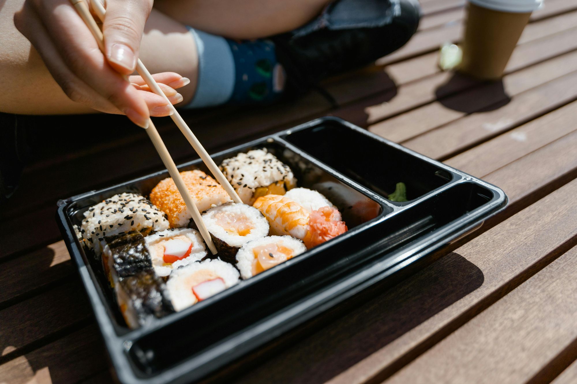 Osoba jedząca sushi z pudełka na stole z drewna, używająca pałeczek.