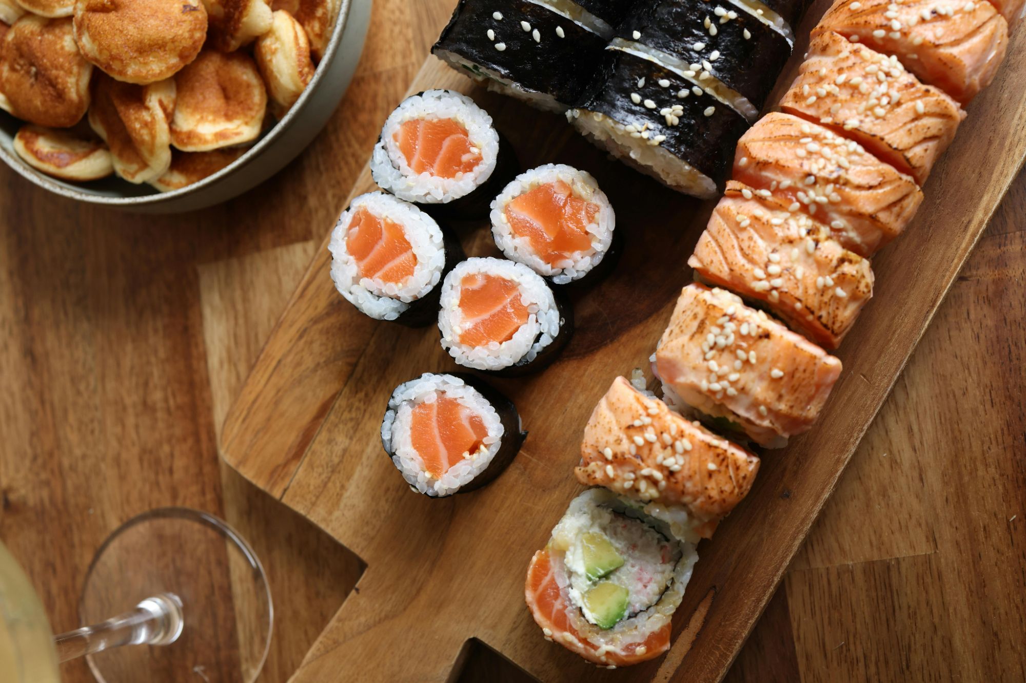 Sushi na drewnianej desce: rolki z łososiem, awokado oraz chrupiące ciasteczka w tle.