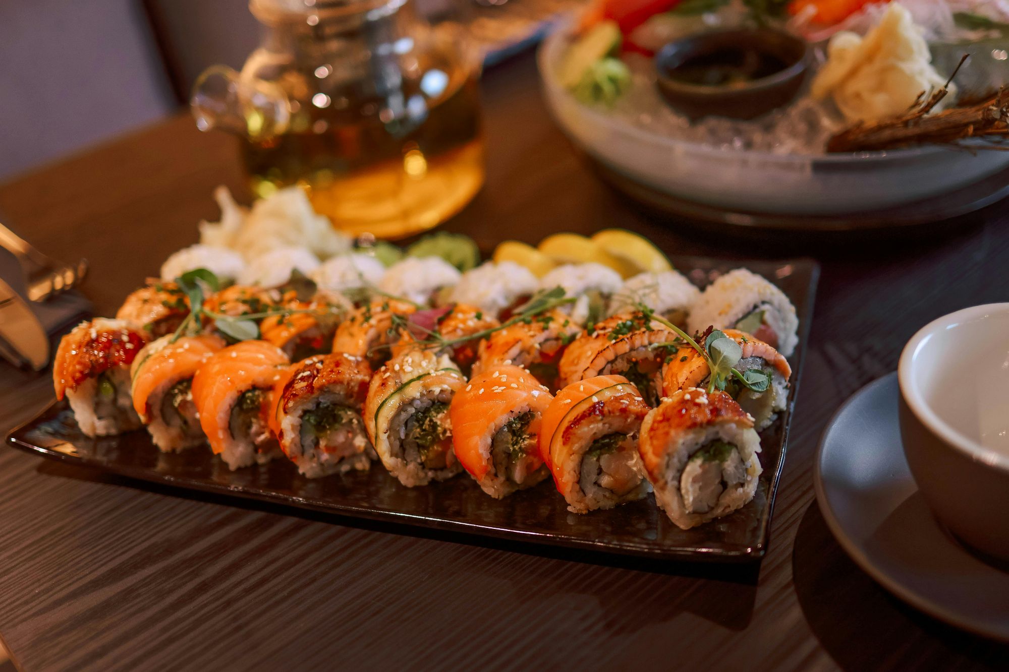 Taca z różnorodnym sushi, w tym rolki z łososiem, awokado i innymi składnikami na tle zielonej herbaty.