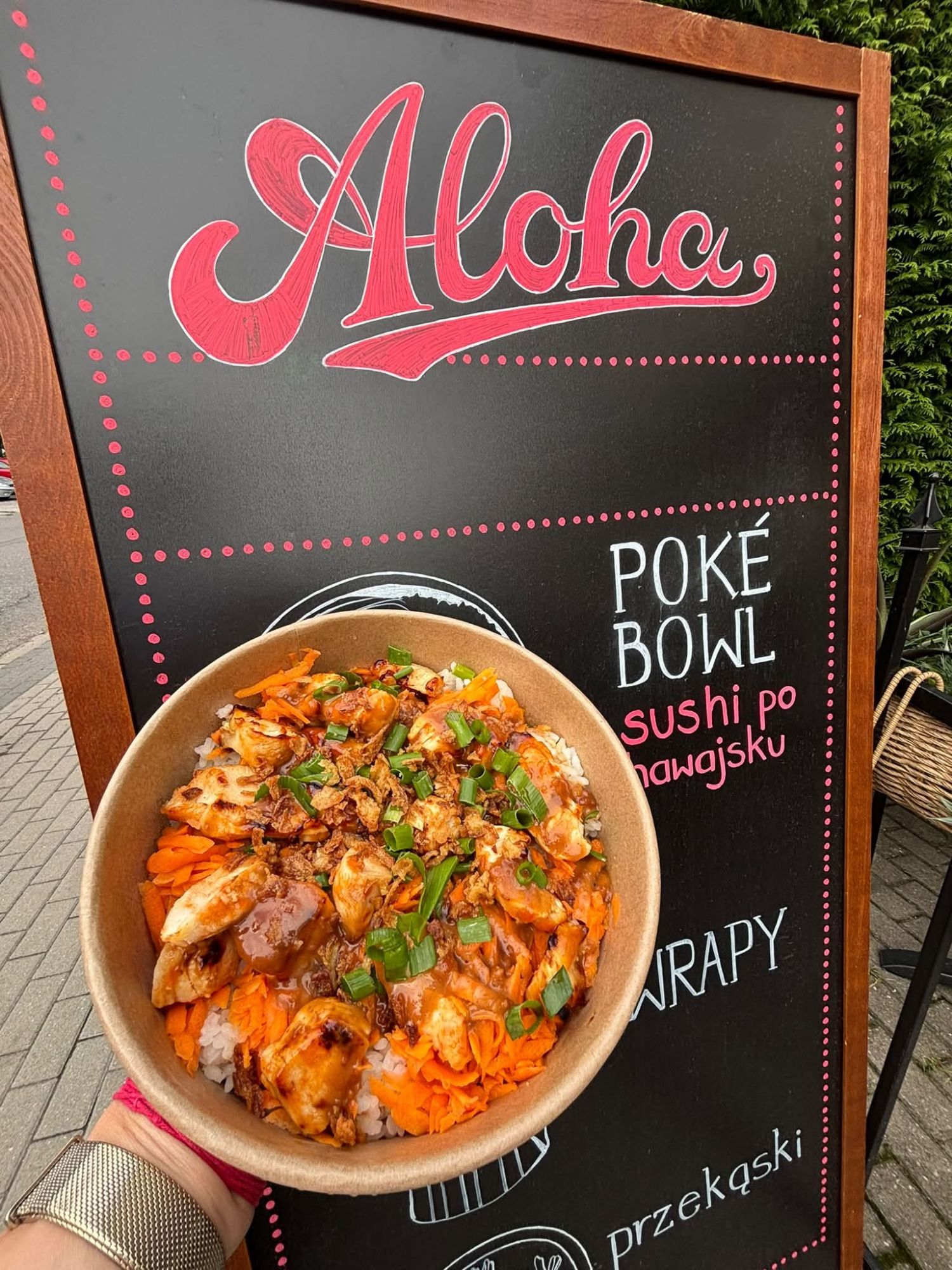 Miska poké z ryżem, warzywami i grillowanym kurczakiem, trzymana przed tablicą z napisem "Aloha".