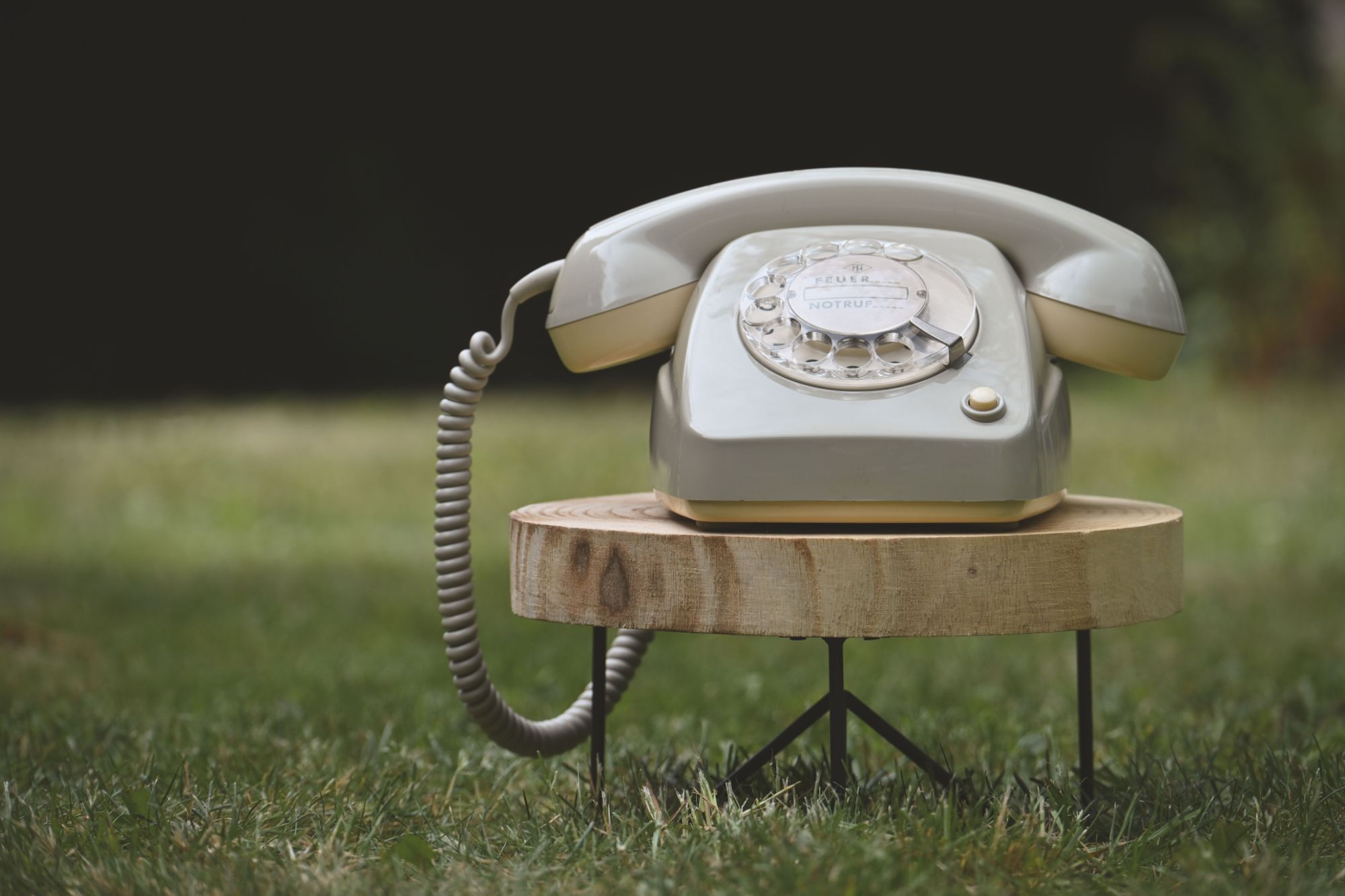 Stary telefon stacjonarny na drewnianym stojaku na trawie, w otoczeniu zieleni.