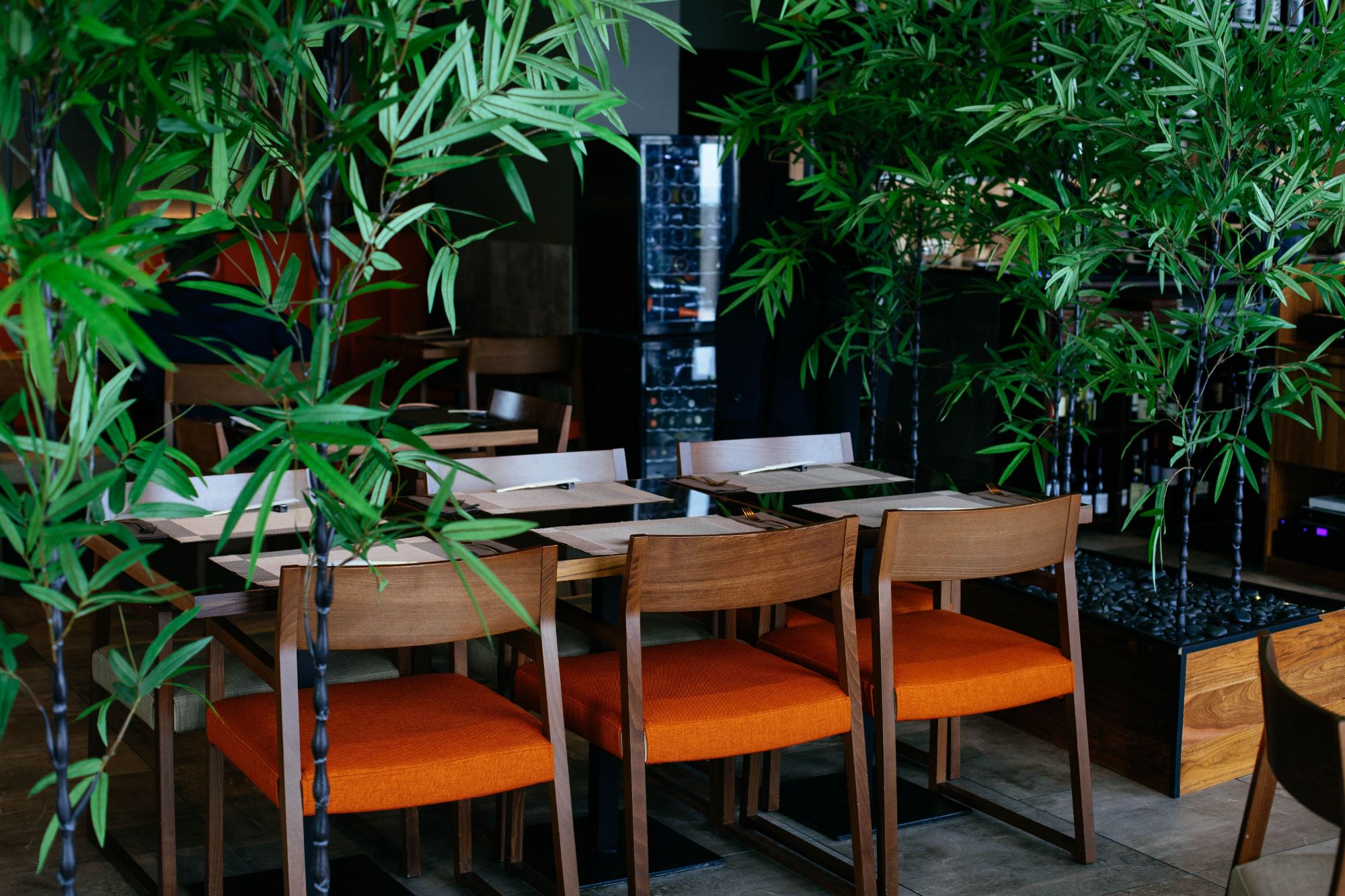 Nowoczesna restauracja z drewnianymi stołami i pomarańczowymi krzesłami, otoczona zielonymi roślinami bambusa.