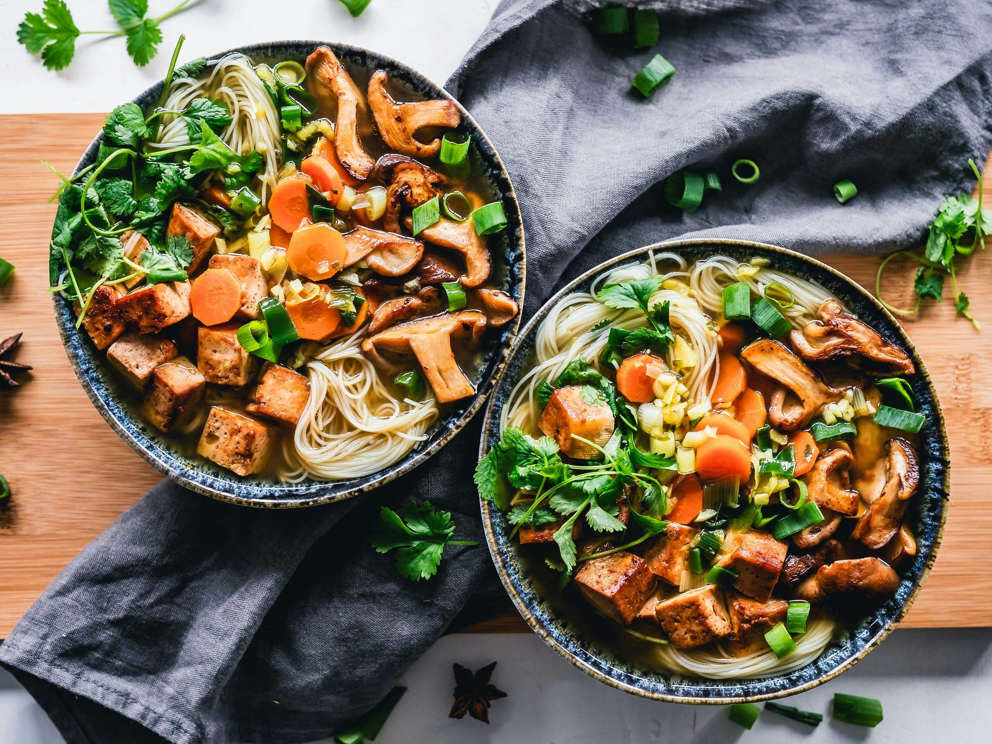 Dwie miski z zupą ramen z makaronem, tofu, warzywami i ziołami na drewnianej desce.