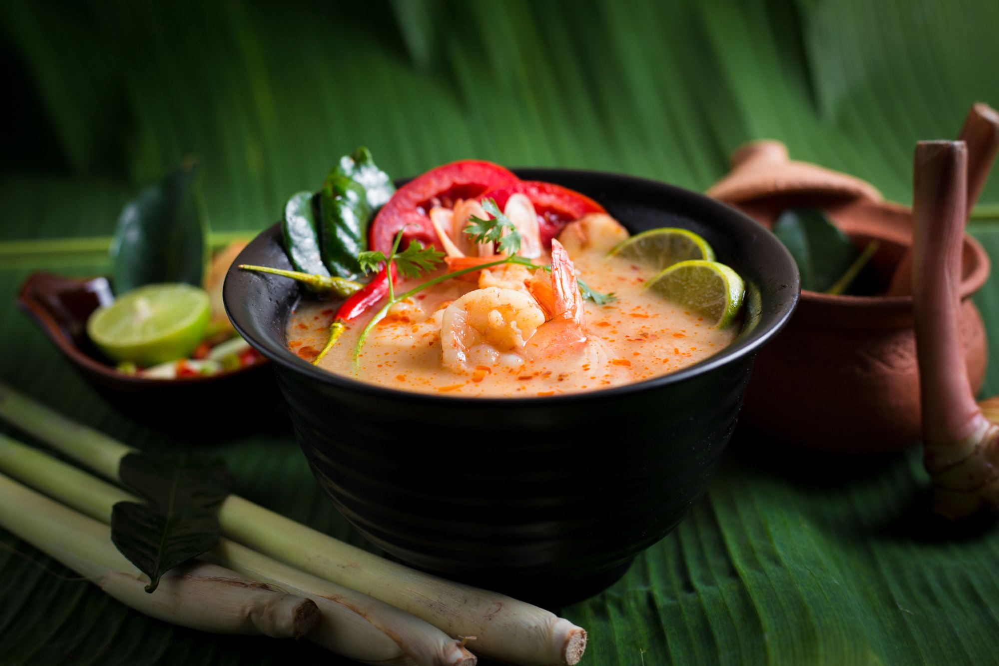 Bowl z zupą tajską Tom Yum, krewetki, zioła, pomidory i limonka na tropikalnym tle.