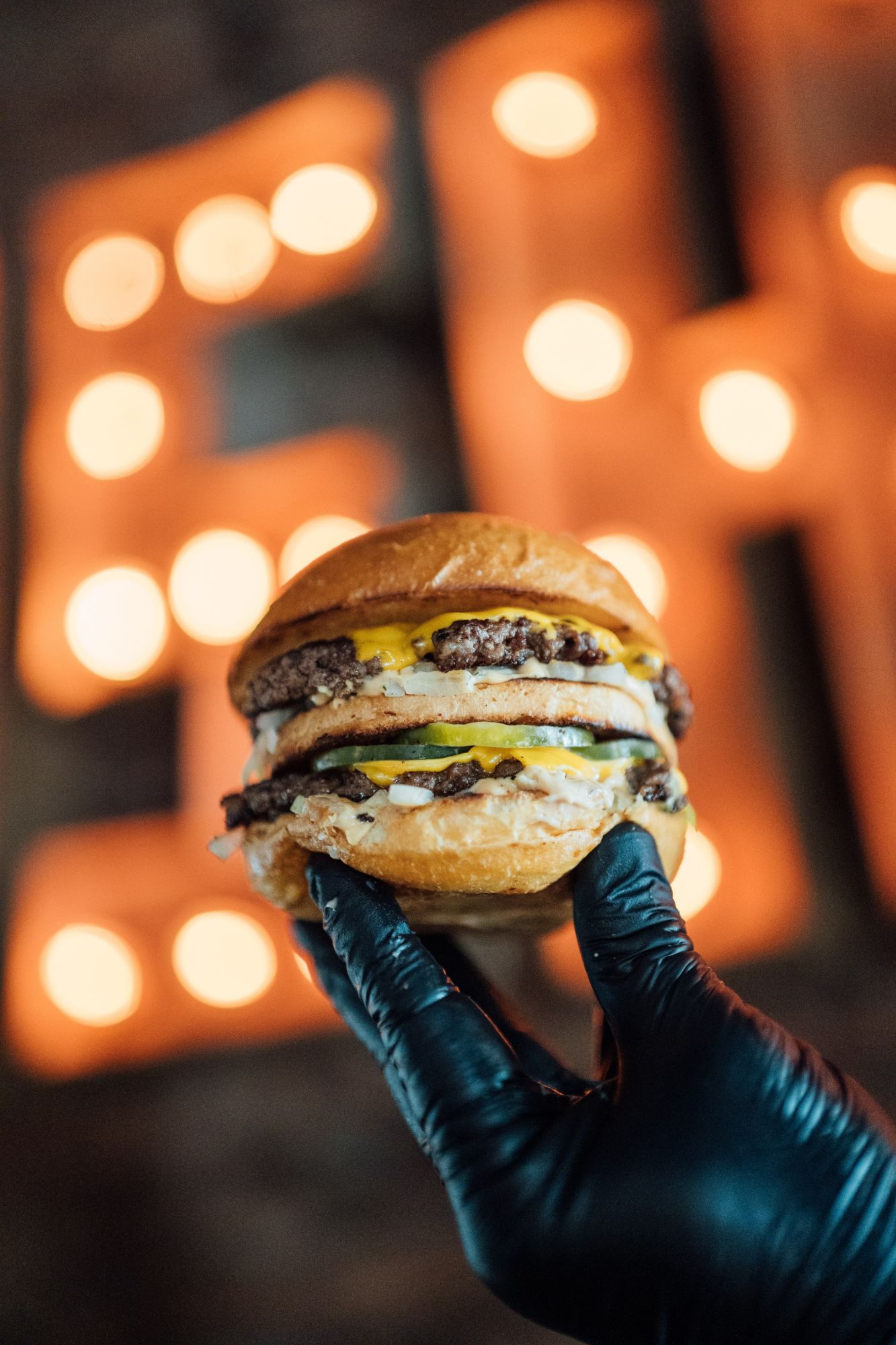 Soczysty burger z podwójnym mięsem, serem i majonezem, trzymany w rękawiczce, w tle neonowe światła.