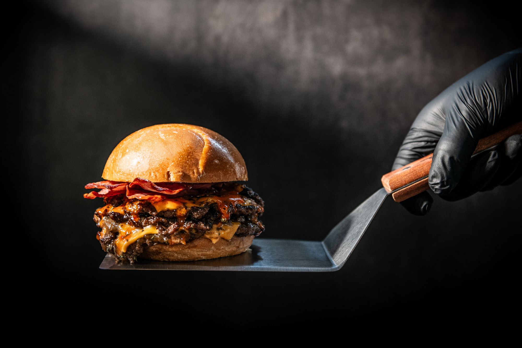 Soczysty burger z serem i boczkiem, trzymany w rękawicy, na stalowej łopatce z drewnianym uchwytem.