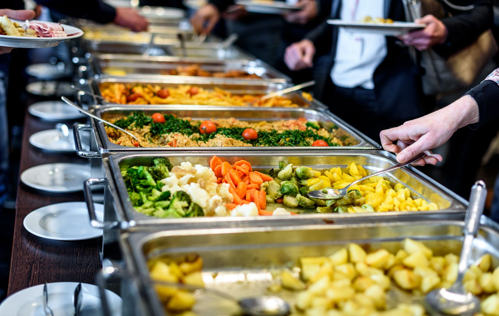 Bufet z daniami obiadowymi, w tym warzywami i ryżem, z gośćmi serwującymi jedzenie na talerze.