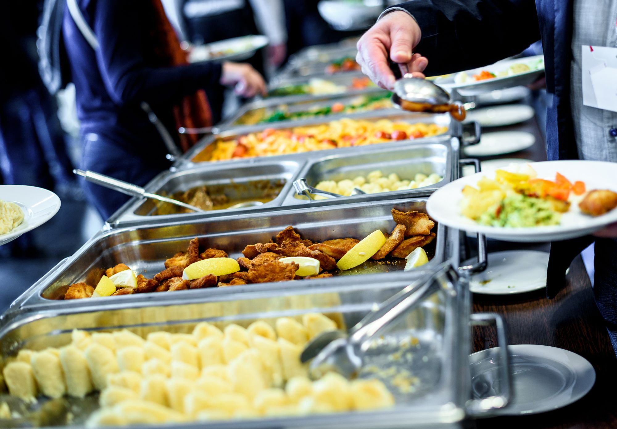 Bufet z różnorodnymi potrawami: pierogi, sałatki, dania główne i owoce. Osoba nakłada jedzenie na talerz.