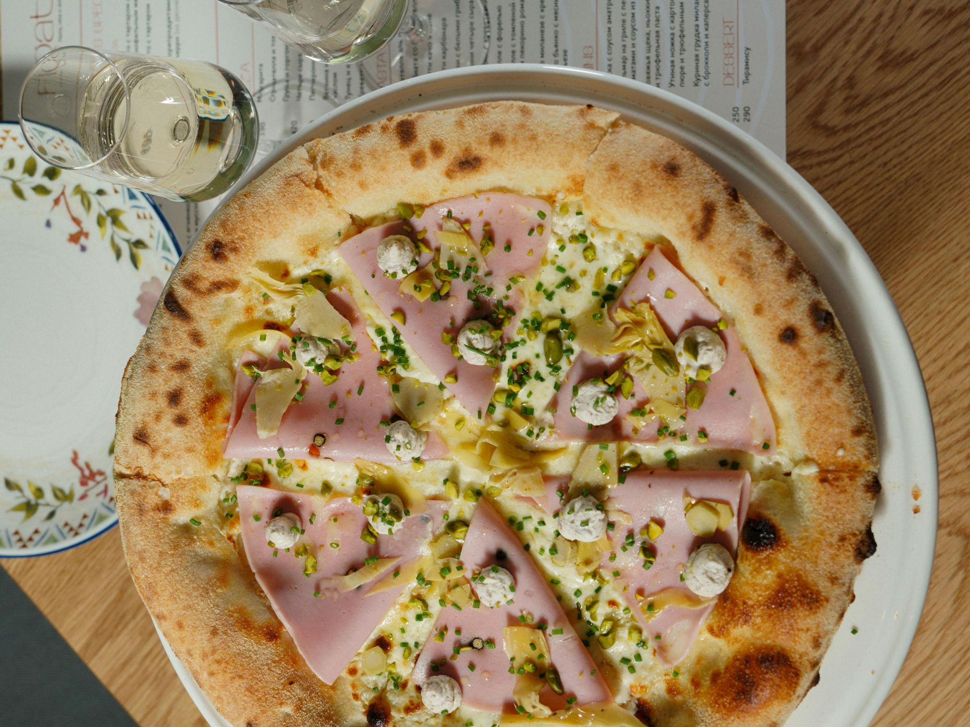 Pizza z szynką, serem, karczochami i szczypiorkiem, podana na talerzu z białym winem w tle.