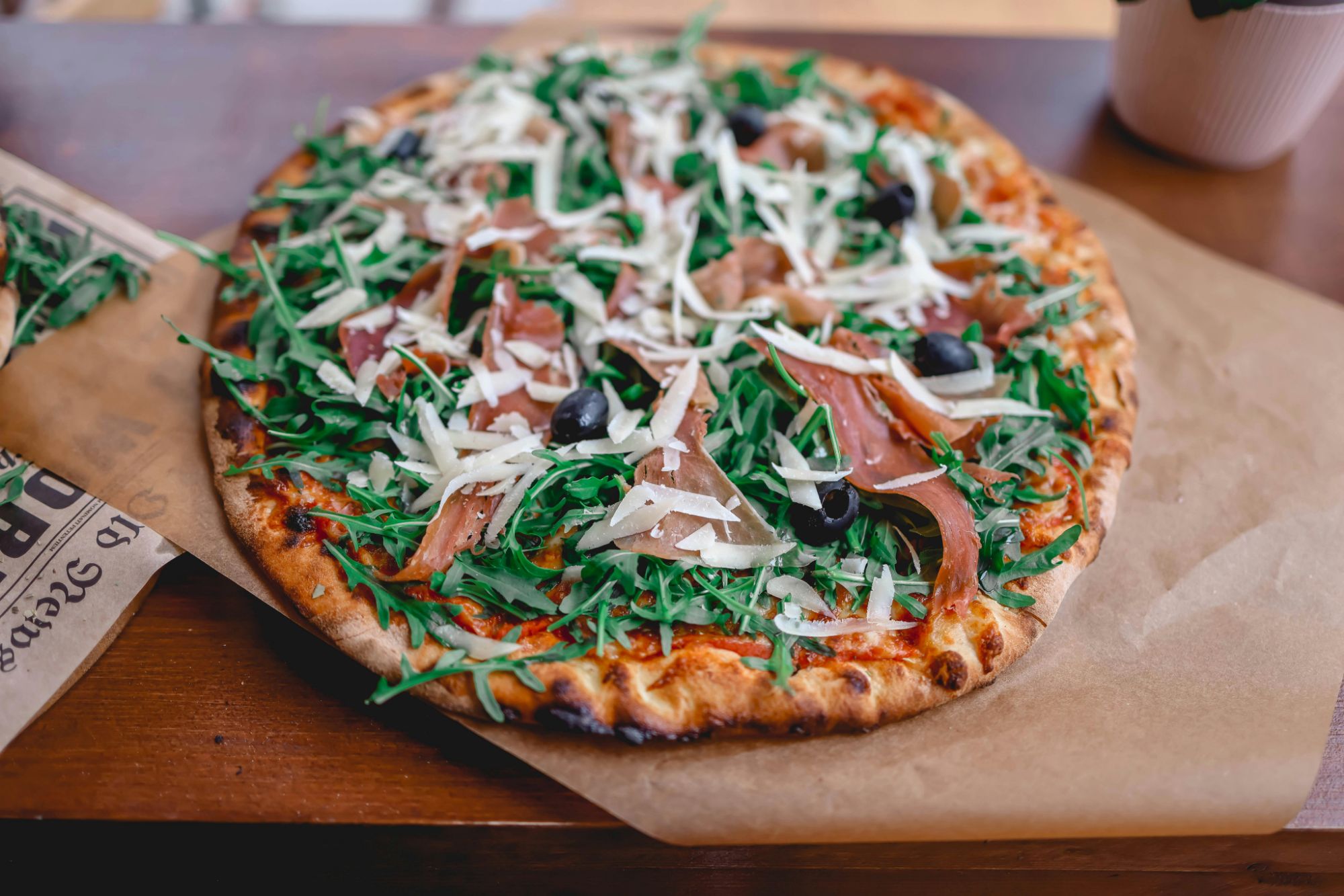Pizza s rukolom, pršutom, maslinama i sirom, poslužena na drvenom stolu.
