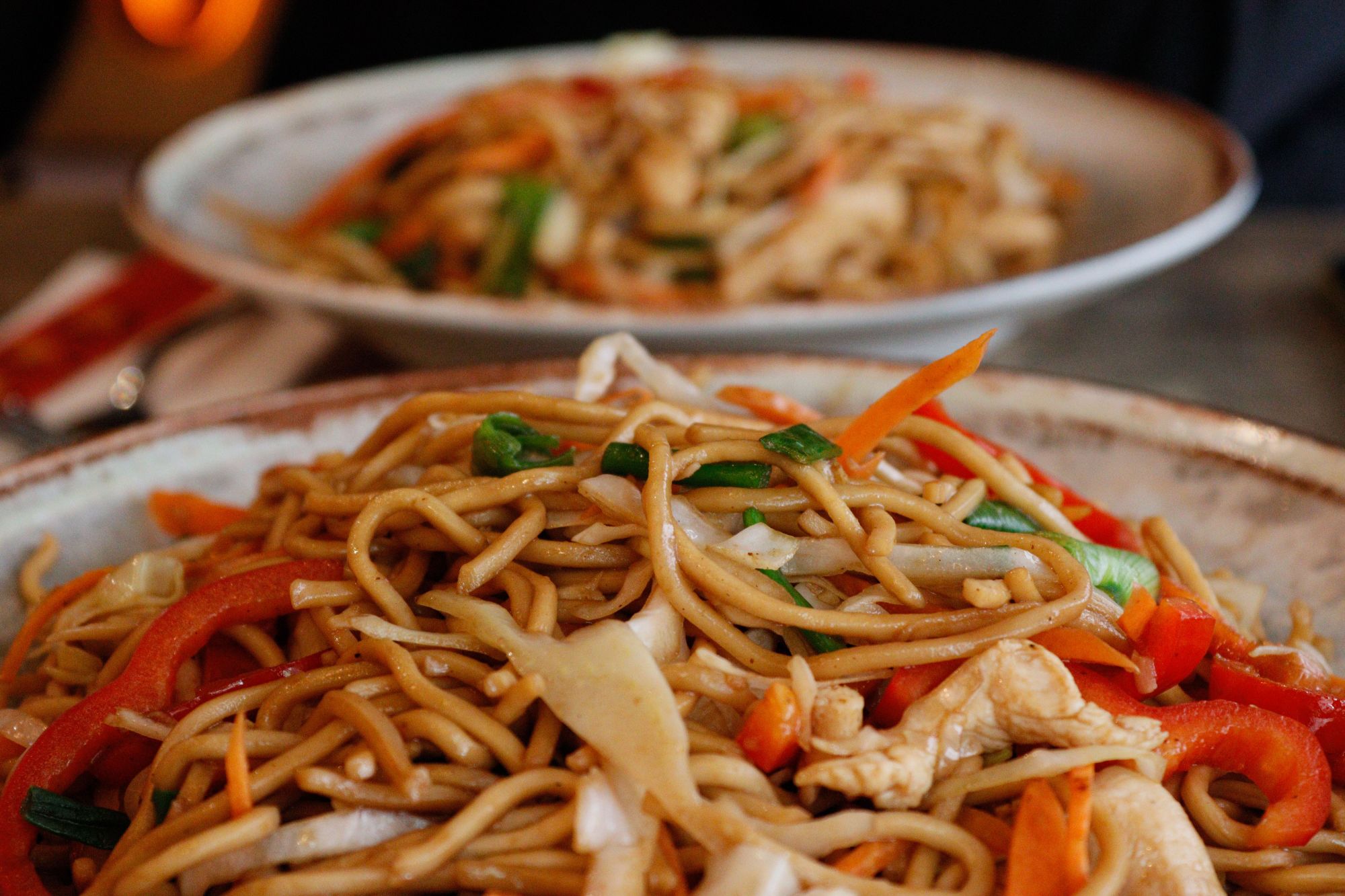 Makaron chow mein z warzywami i kurczakiem w tłem talerza z podobnym daniem.