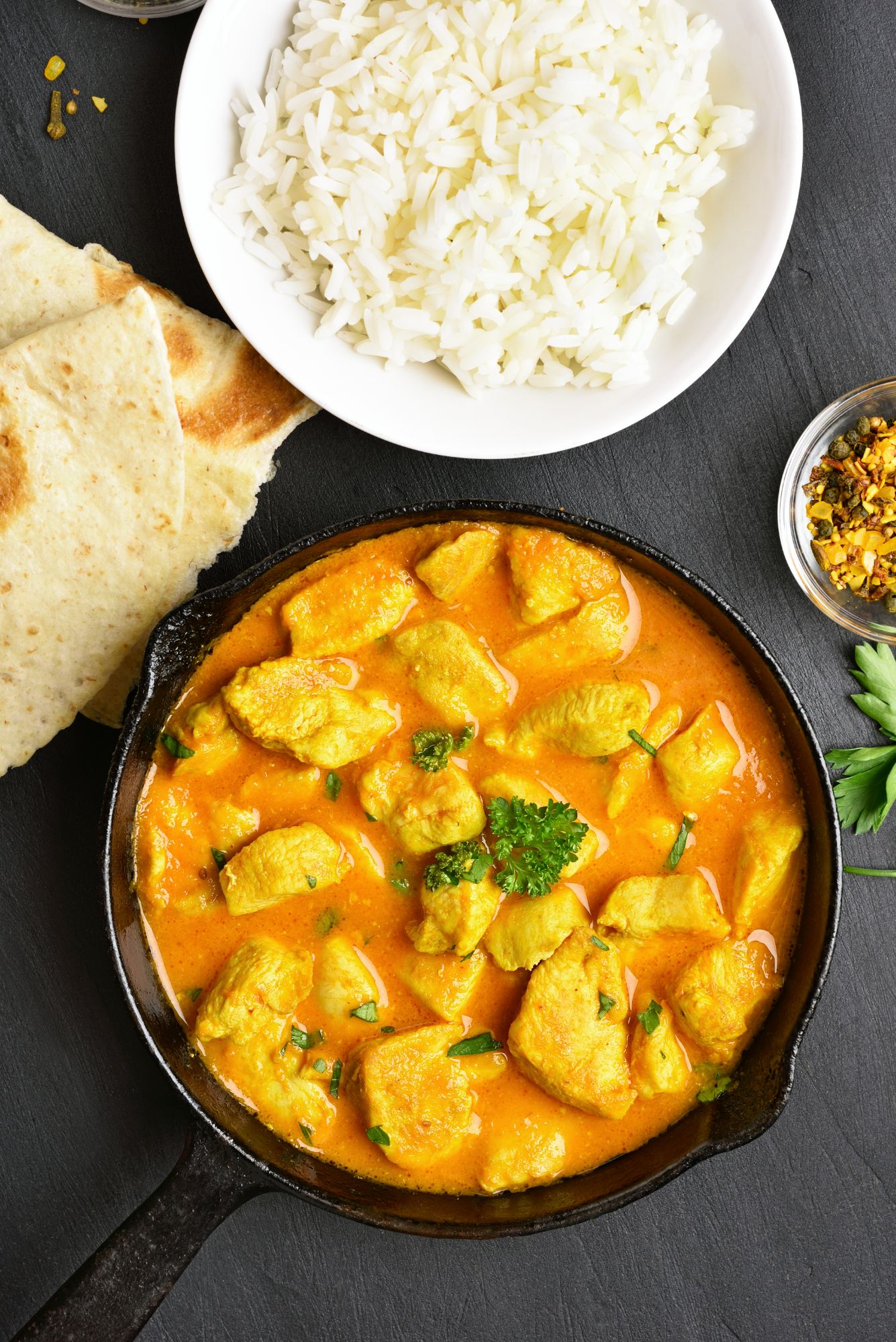 Potrawa z kurczaka w sosie curry, biały ryż, chleb naan oraz przyprawy na czarnej powierzchni.