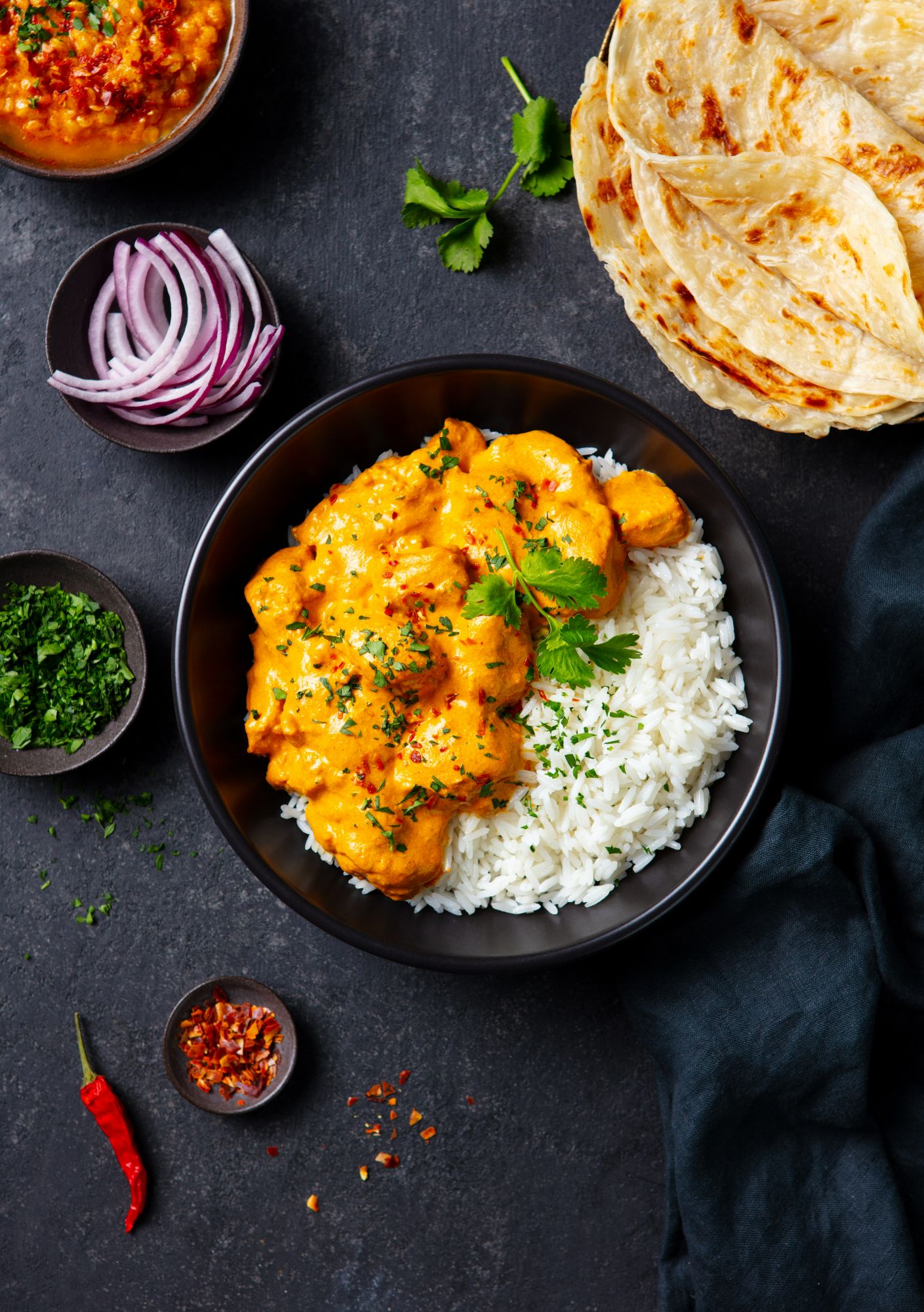 Kremowe curry z kurczakiem podane z ryżem, cebulą, kolendrą i chlebem naan na ciemnym tle.