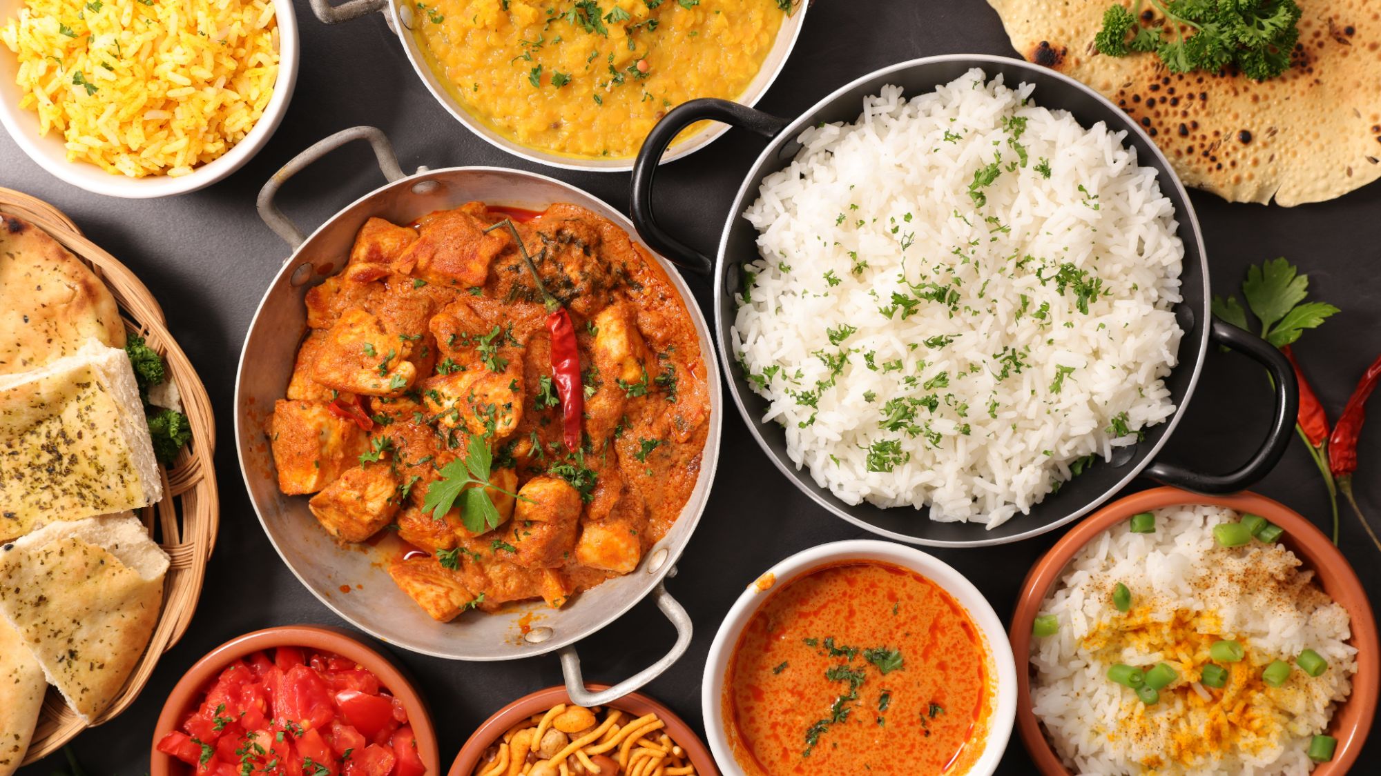 Różnorodne dania indyjskie: ryż, curry, naan, sałatka i soczewica w jedzeniu na czarnym tle.