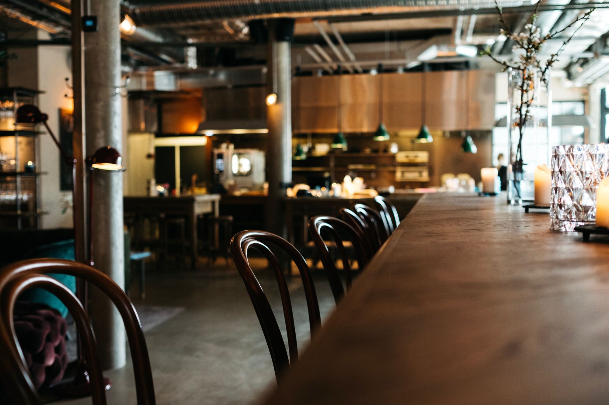 Moderno uređen restoran s drvenim barom, stolicama i kuhinjom u pozadini. Prirodna svjetlost i industrijski elementi.