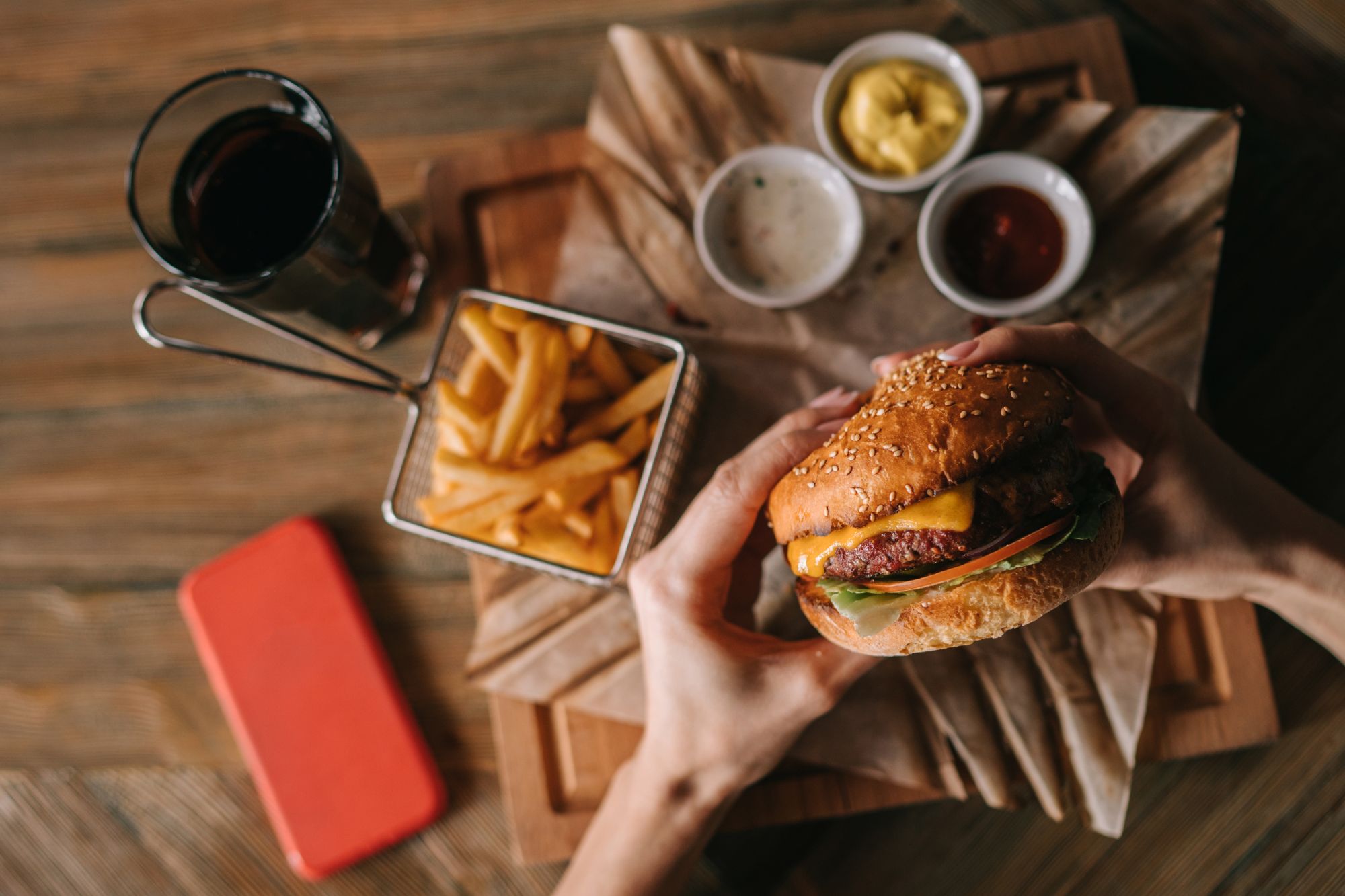 Osoba drži hamburger s cheddar sirom, uz pomfrit i umake na drvenom stolu.