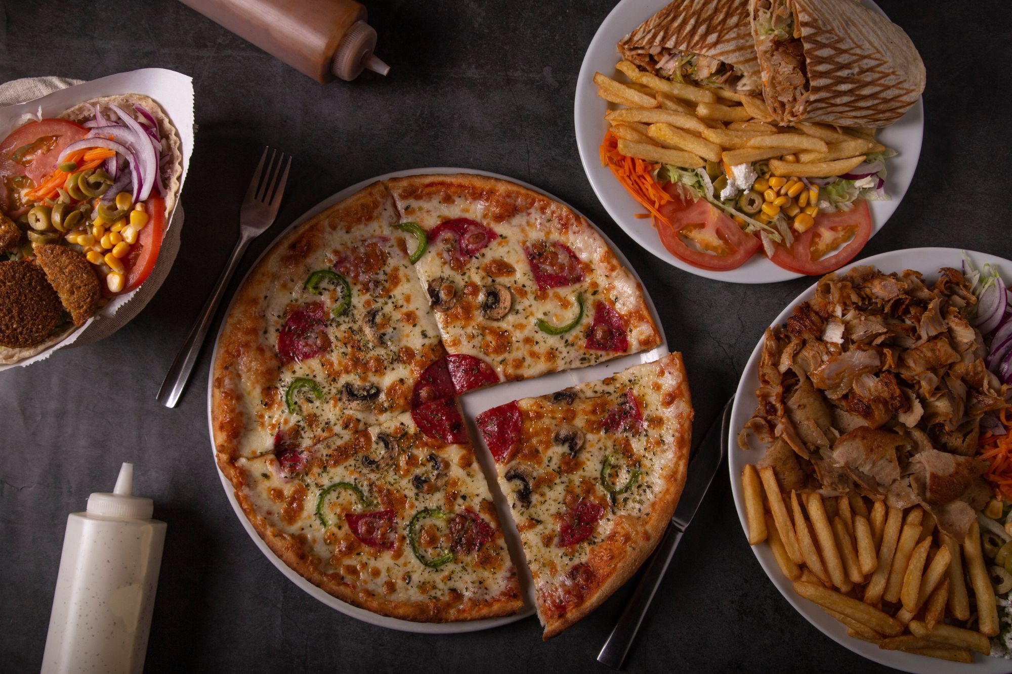 Pizza z pepperoni, papryką i oliwkami, talerze z kebabem, frytkami oraz sałatką, na ciemnym stole.