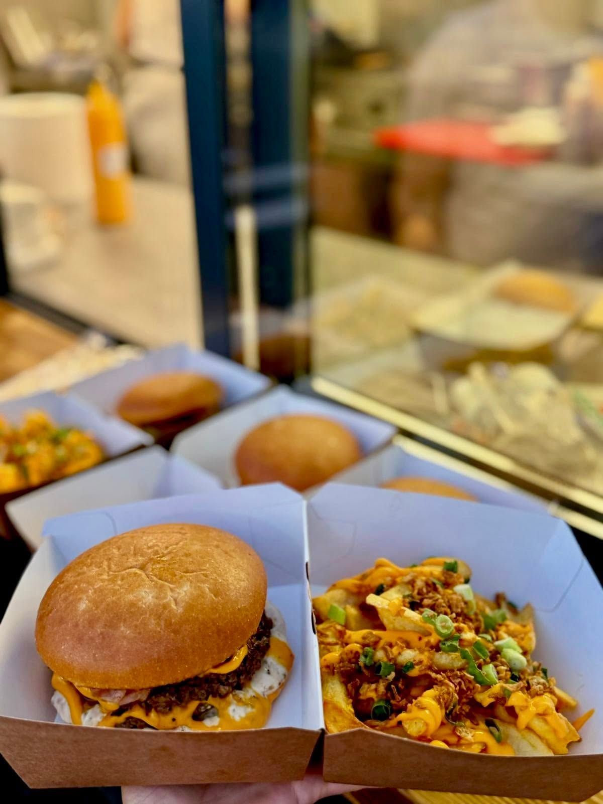 Burger cu brânză și cartofi prăjiți, servite într-un coș din carton, cu un fundal de bucătărie aglomerată.