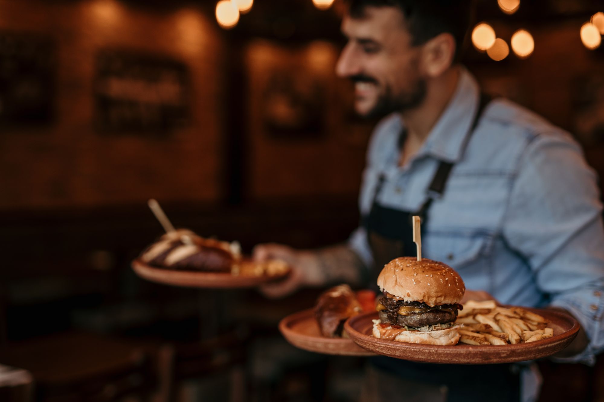 Un chelner zâmbitor aduce două platouri cu burger și cartofi prăjiți într-un restaurant cu un ambient cozy.