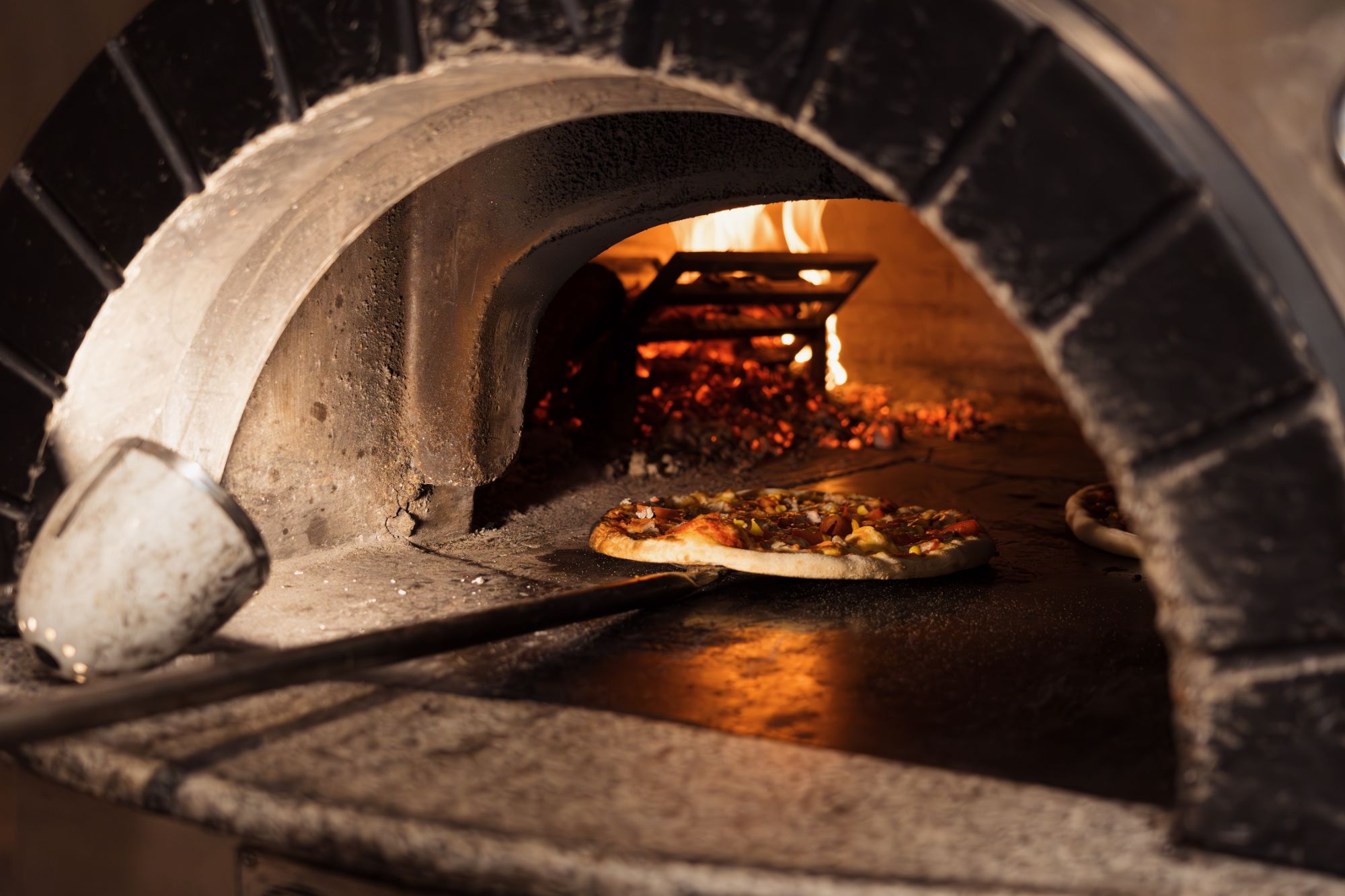 Pizzę wypiekająca w piecu opalanym drewnem, z widocznym ogniem i popiołem.