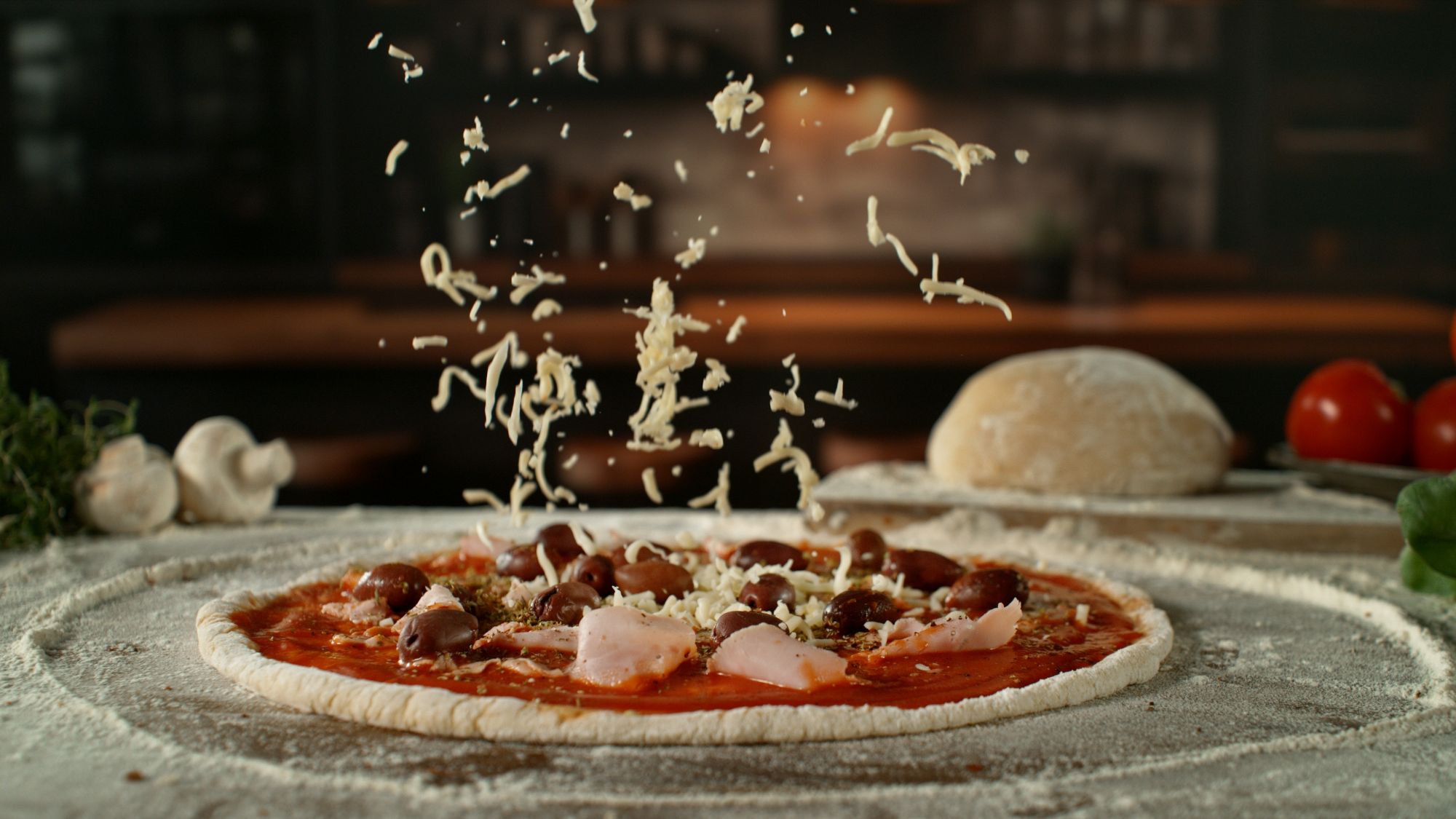 Ser pokrywa pizzę z dodatkami na przygotowanej cieście, w tle widać składniki i stół kuchenny.