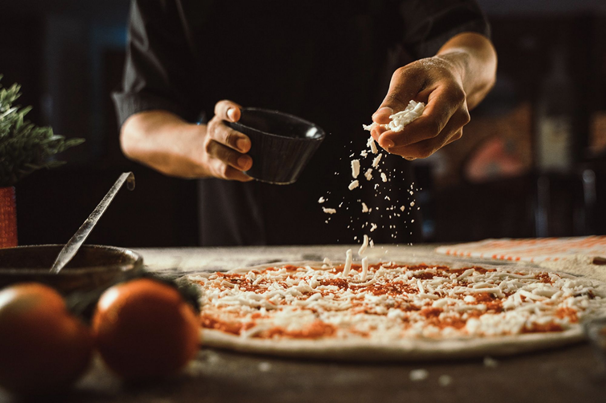 Osoba posypująca serem pizzę w restauracji, z pomidorami w tle.