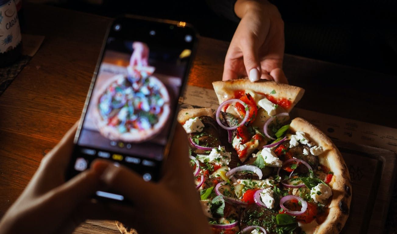 Osoba trzymająca kawałek pizzy z warzywami i serem, obok telefon zrobiony zdjęcie całej pizzy.
