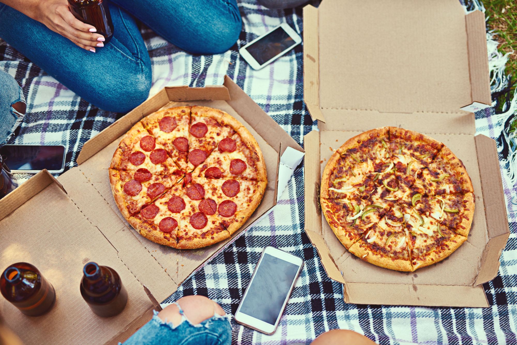 Dwa pudełka pizzy na kocu, w tle kilka smartfonów i butelek piwa, idealne na piknik.
