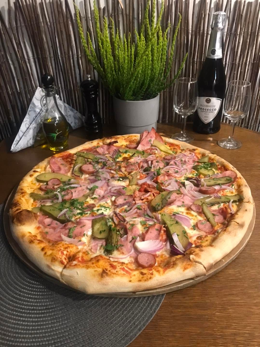 Pizza z warzywami, szynką i serem, podana na stole z winem i kieliszkami w tle.