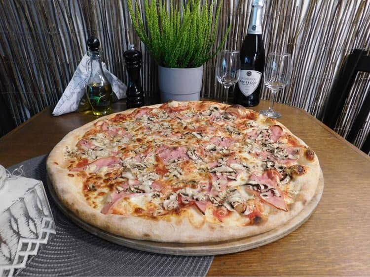Pizza na dużym cieście z szynką, pieczarkami i serem, podana na drewnianym stole z kieliszkami i butelką szampana.