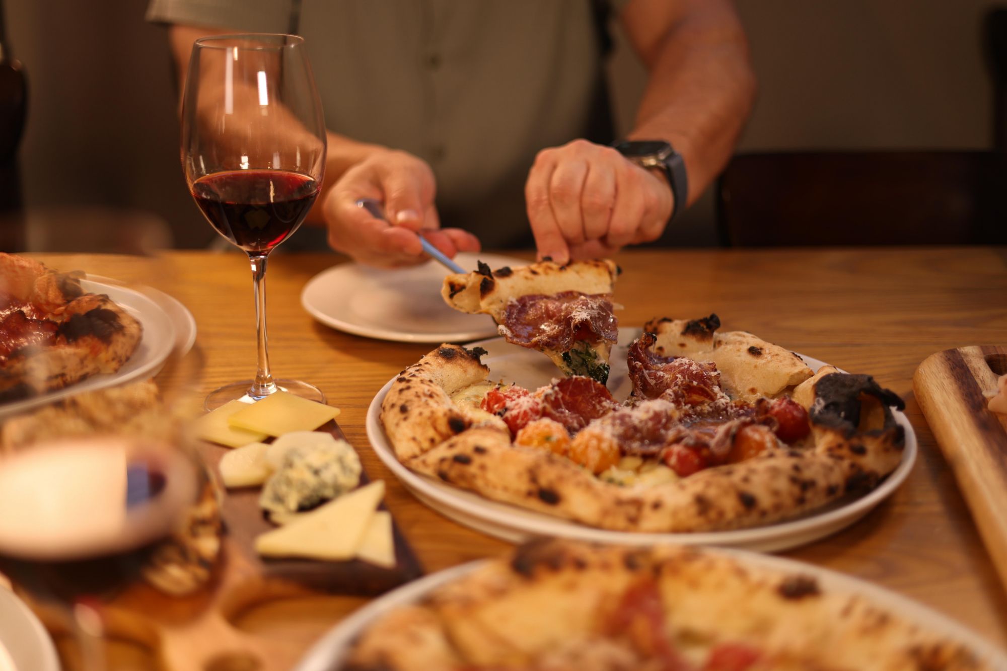 Mężczyzna kroi pizzę na talerzu, obok kieliszek czerwonego wina i deska serów na drewnianym stole.
