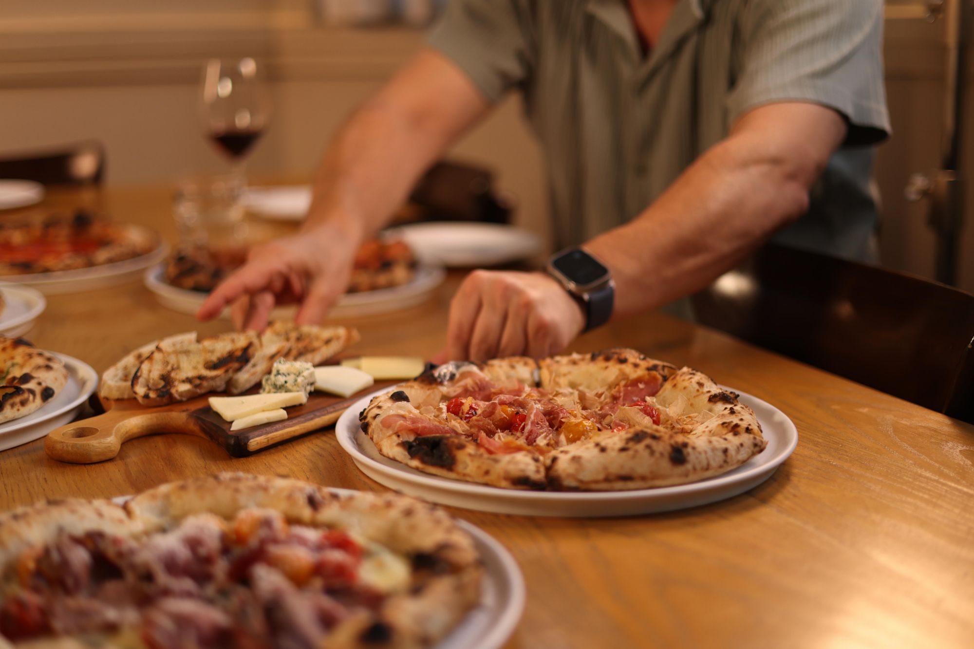 Osoba sięga po pizzę na stole, obok talerza z serem i kieliszkiem wina. W tle widać inne pizze.