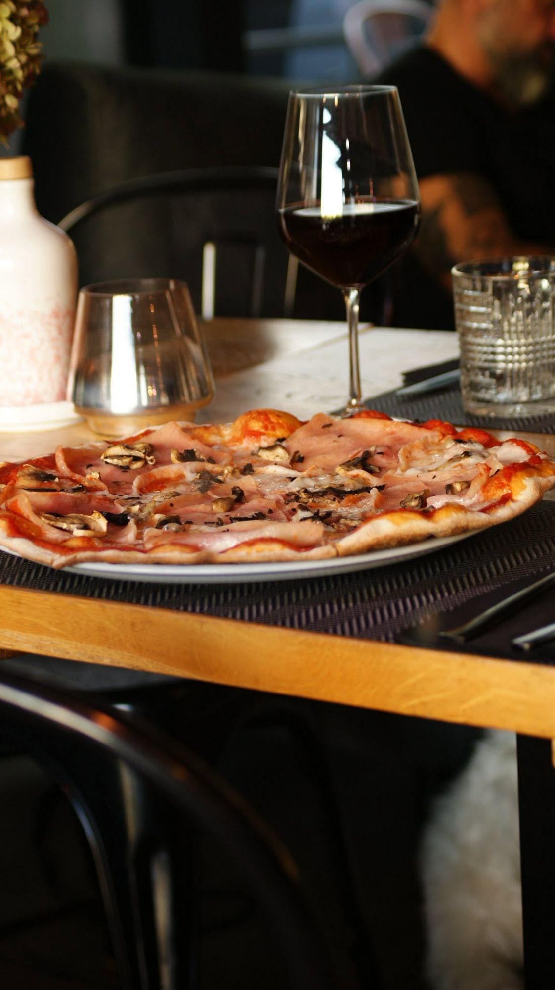 Pizza z szynką i grzybami na stole z winem w tle.