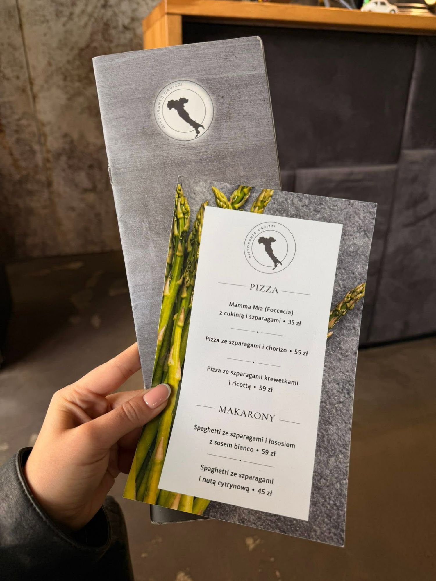Menu of a restaurant featuring pizzas and pasta dishes, held by a person in a casual setting.